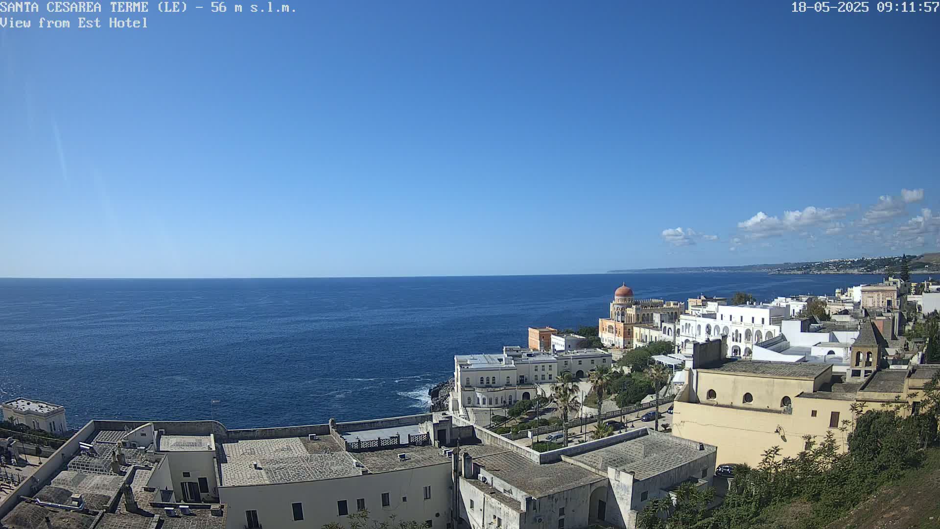 A coastal town with white buildings and a domed structure overlooks a calm blue sea under a mostly clear sky with a few scattered clouds.