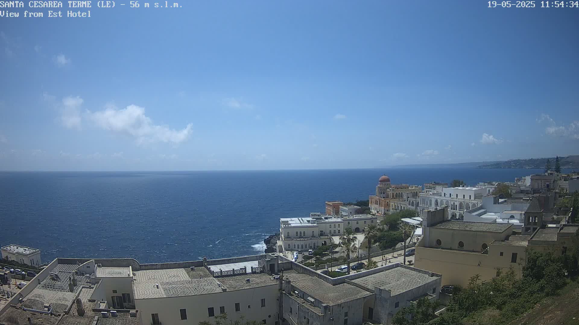 A coastal town with white buildings overlooks a calm, blue sea under a mostly sunny sky with a few scattered clouds.