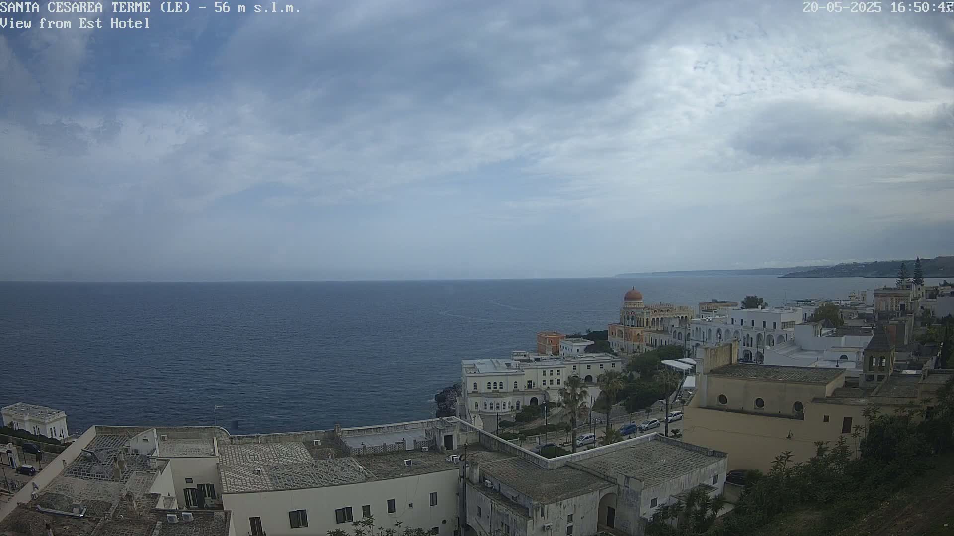 A coastal town with white buildings, including a domed structure, overlooks a calm sea under a partly cloudy sky.