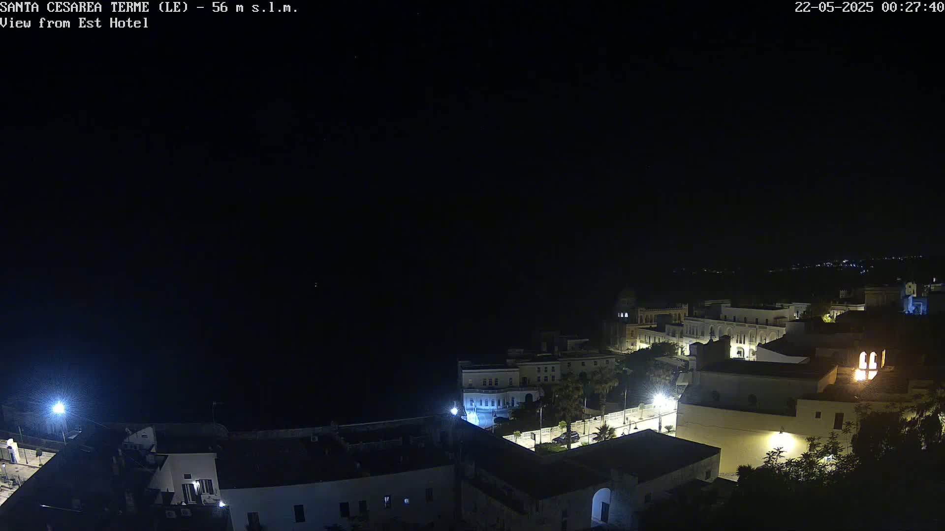 A nighttime, high-angle view of a coastal town with buildings illuminated, overlooking a dark body of water under a clear, starlit sky.