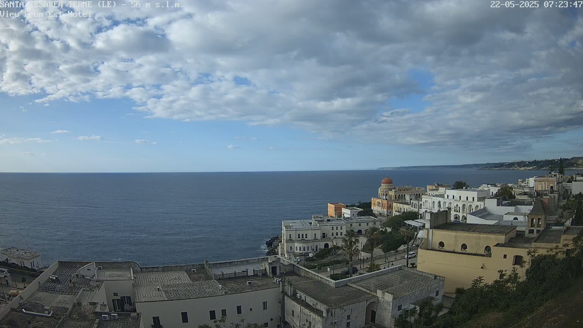 A coastal town with white buildings and a domed structure overlooks a calm sea under a partly cloudy sky.