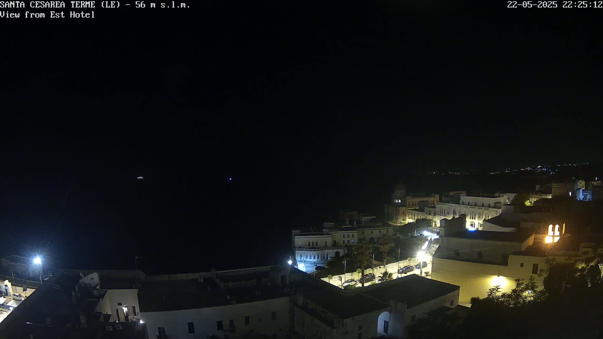 A nighttime, high-angle view of a coastal town with dimly lit buildings and streets, under a dark, clear sky.