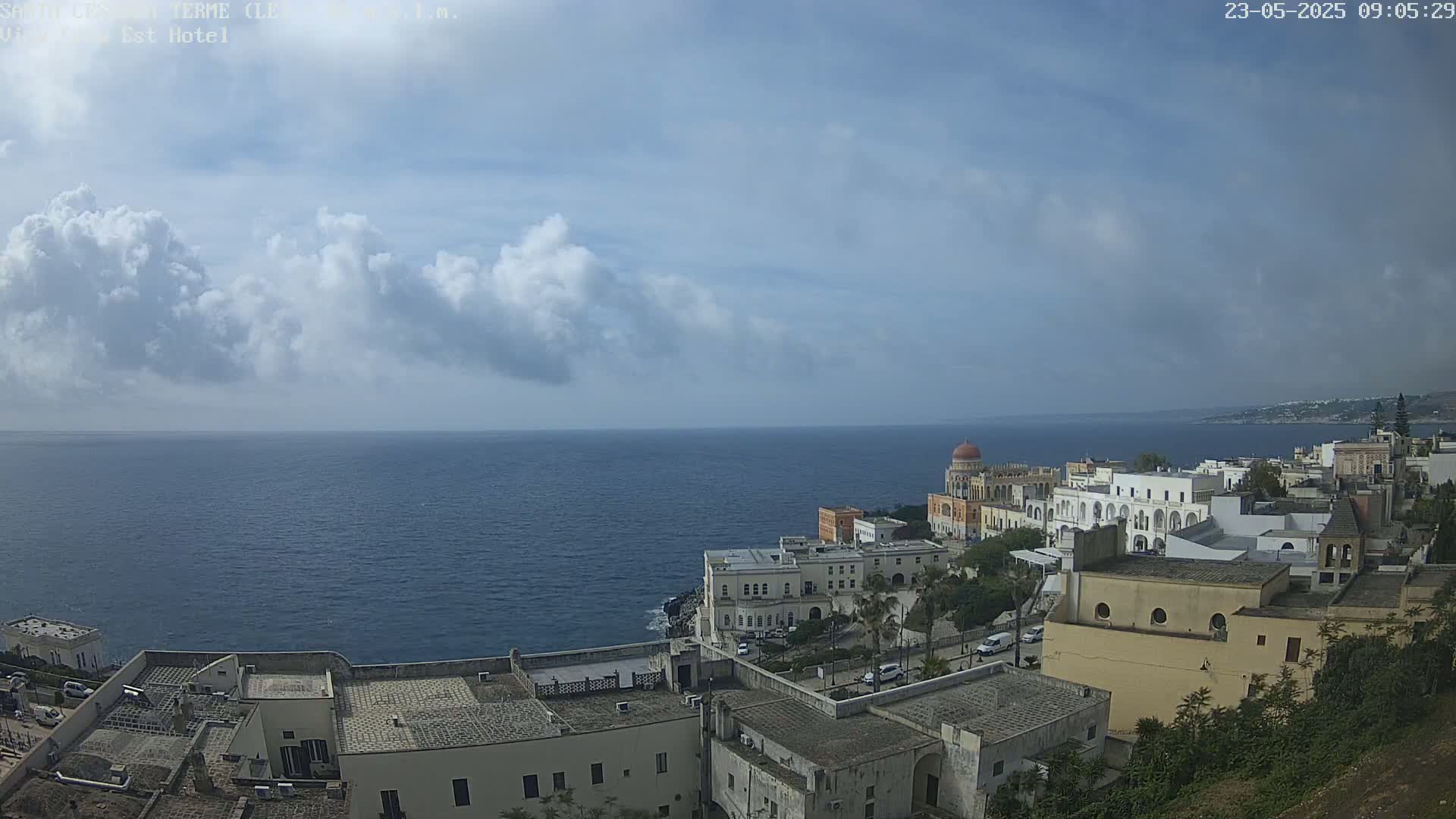 A coastal town with white buildings and a dome-topped structure is visible, overlooking a calm sea under a partly cloudy sky.