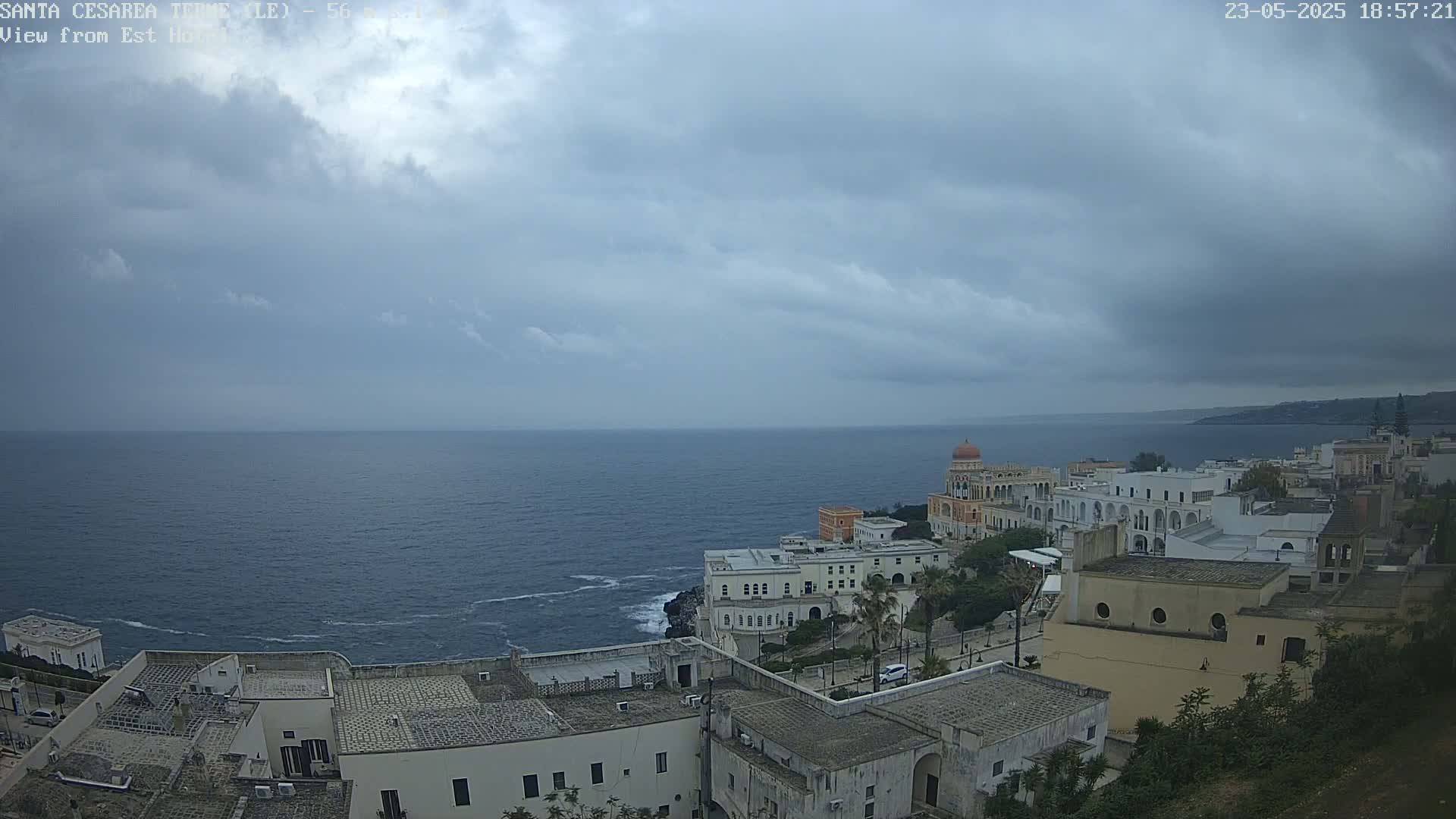 A coastal town with white buildings and a domed structure overlooks a calm sea under a cloudy sky.