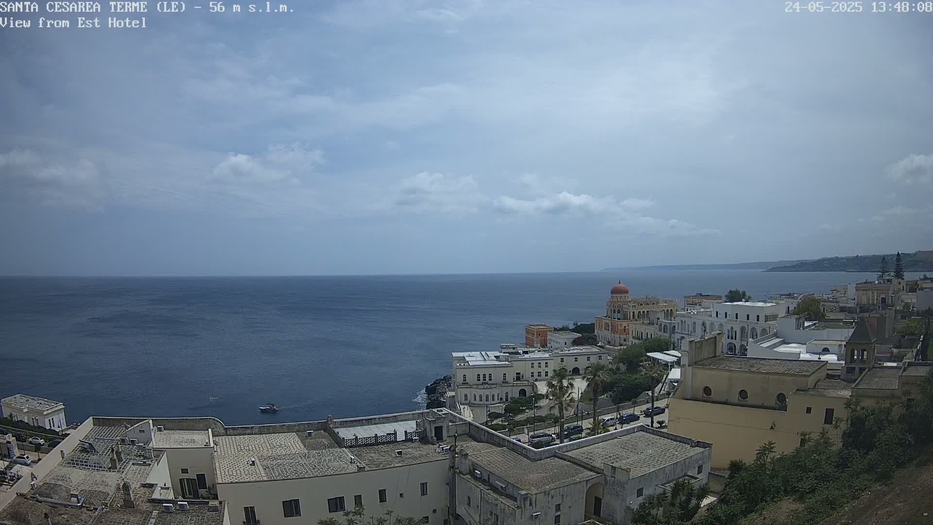 A coastal town with white buildings and a domed structure overlooks a calm sea under a partly cloudy sky.
