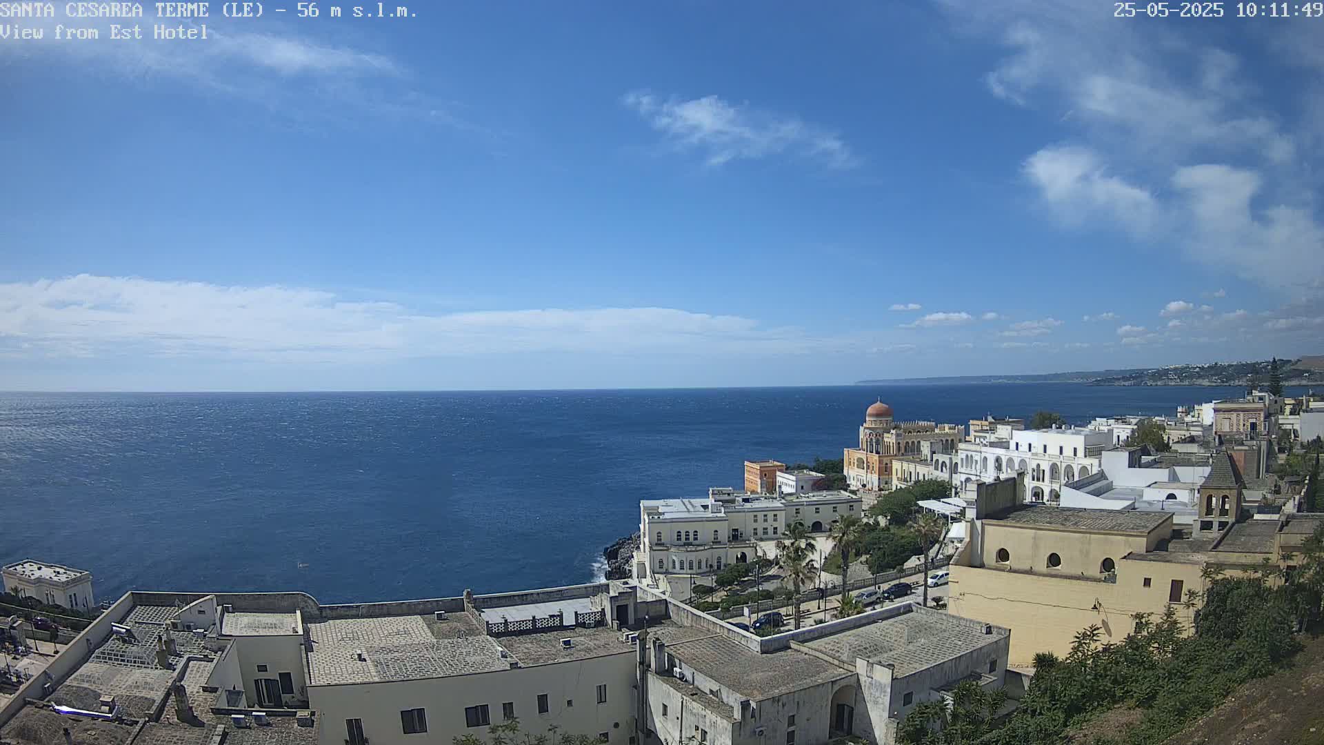 A coastal town with white buildings overlooks a calm blue sea under a mostly sunny sky with a few scattered clouds.