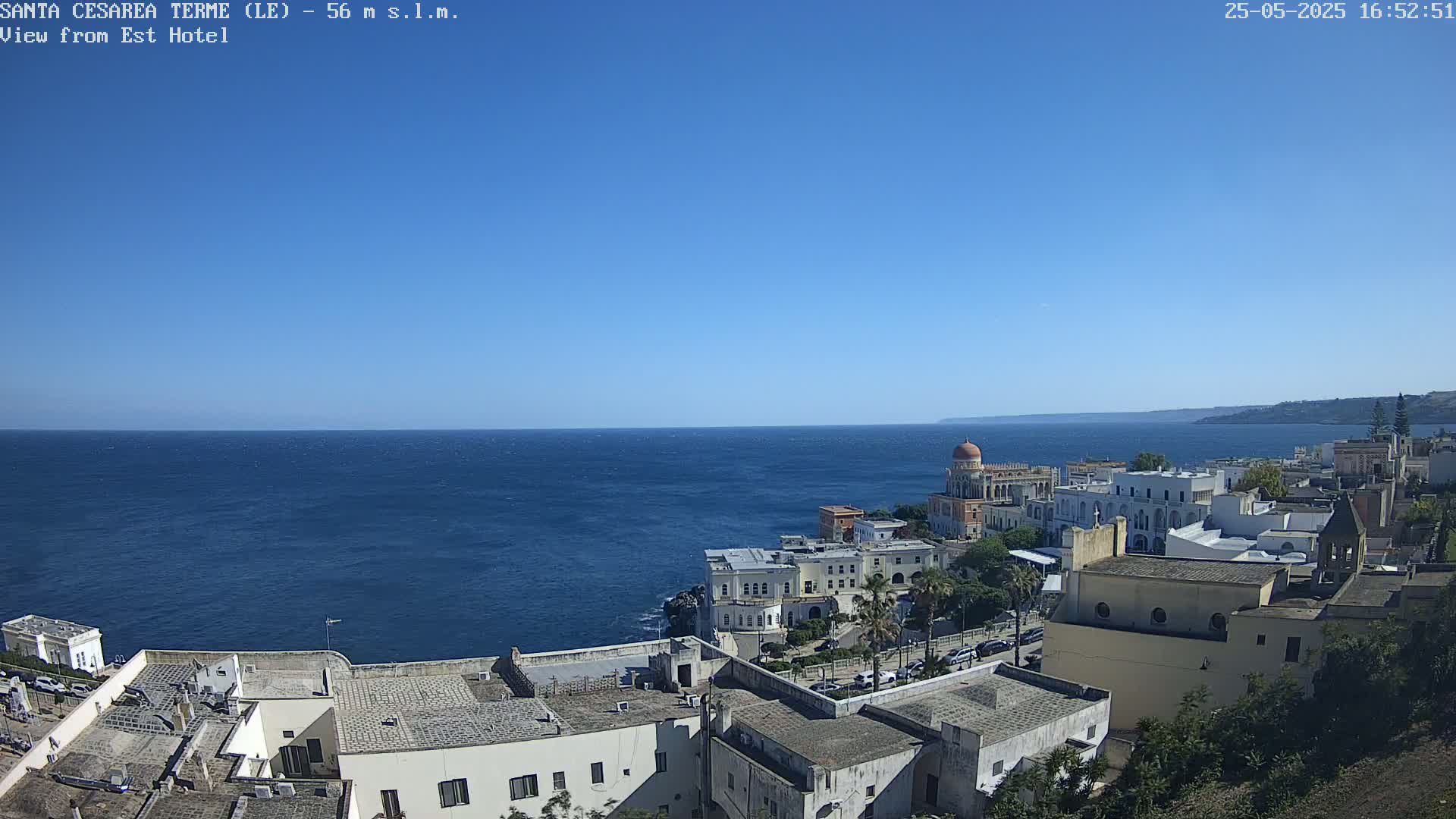 A coastal town with white buildings overlooks a calm, blue sea under a clear, bright blue sky.