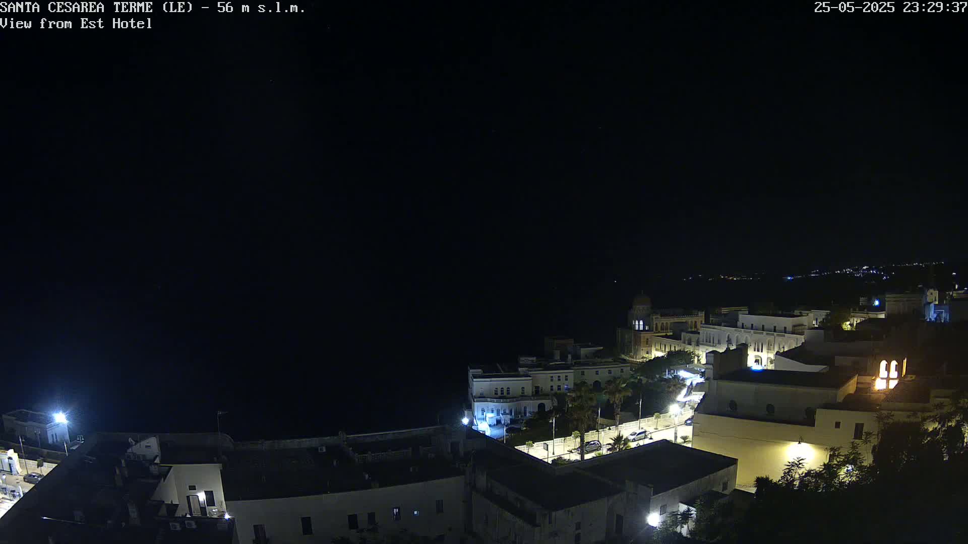 A nighttime, high-angle view of a coastal town with buildings illuminated, overlooking a dark sea under a clear night sky.
