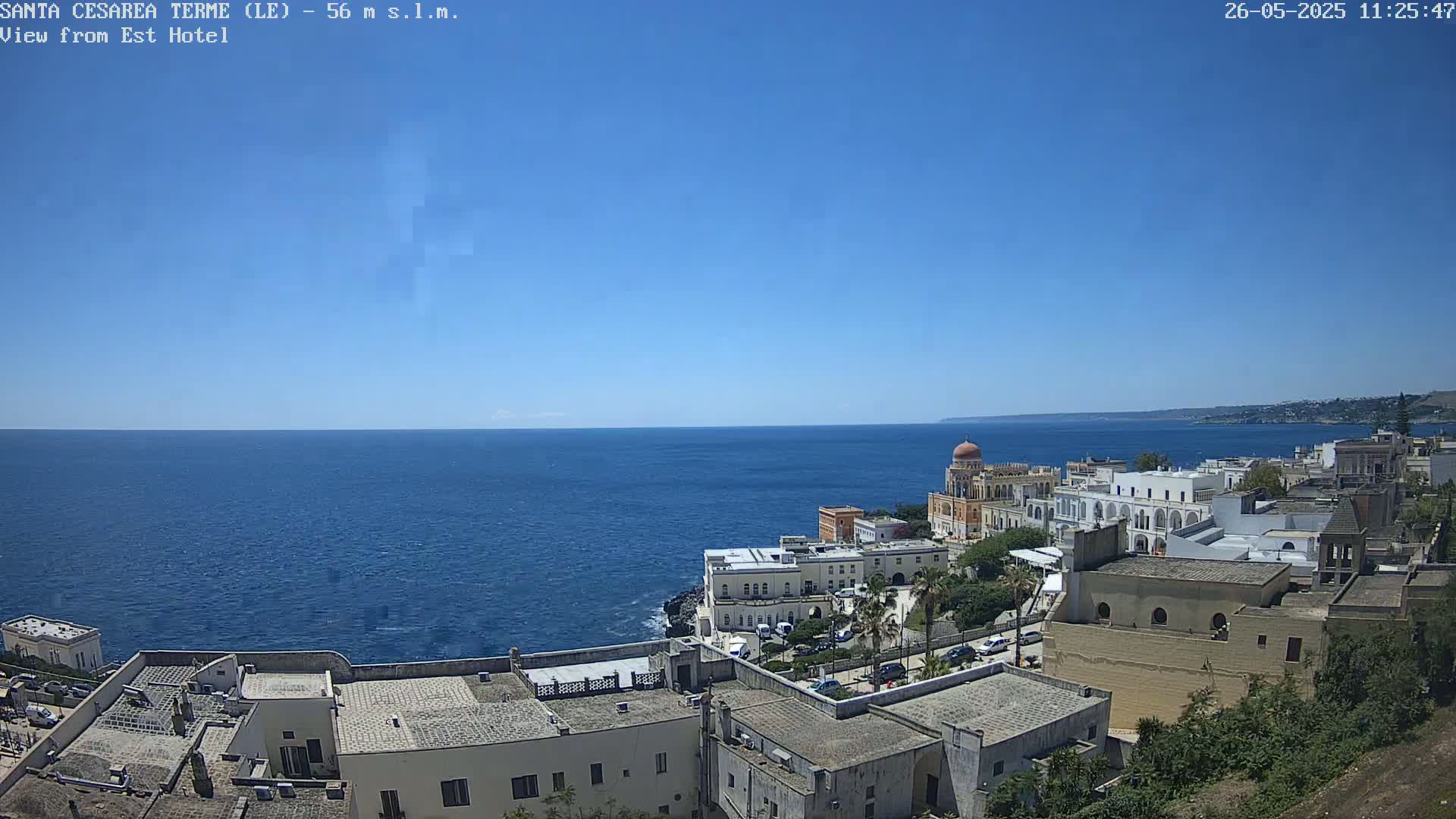 A coastal town with white buildings and a domed structure sits on a cliff overlooking a calm, blue sea under a clear, sunny sky.