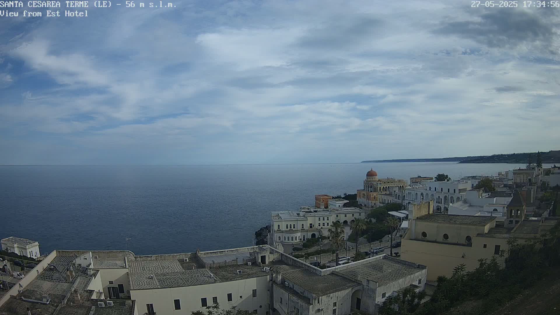 A coastal town with white buildings and a domed structure overlooks a calm sea under a partly cloudy sky.