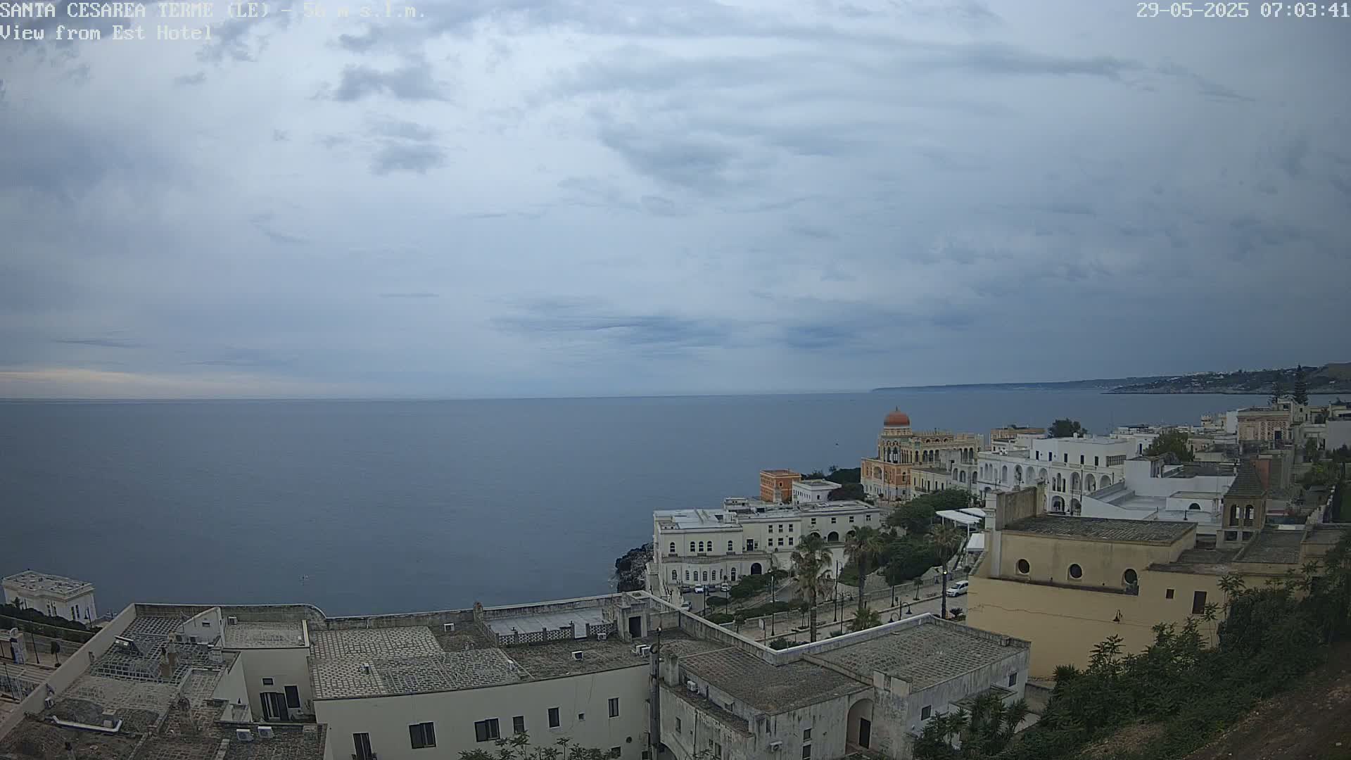 A coastal town with white buildings and a domed structure overlooks a calm sea under a cloudy sky.