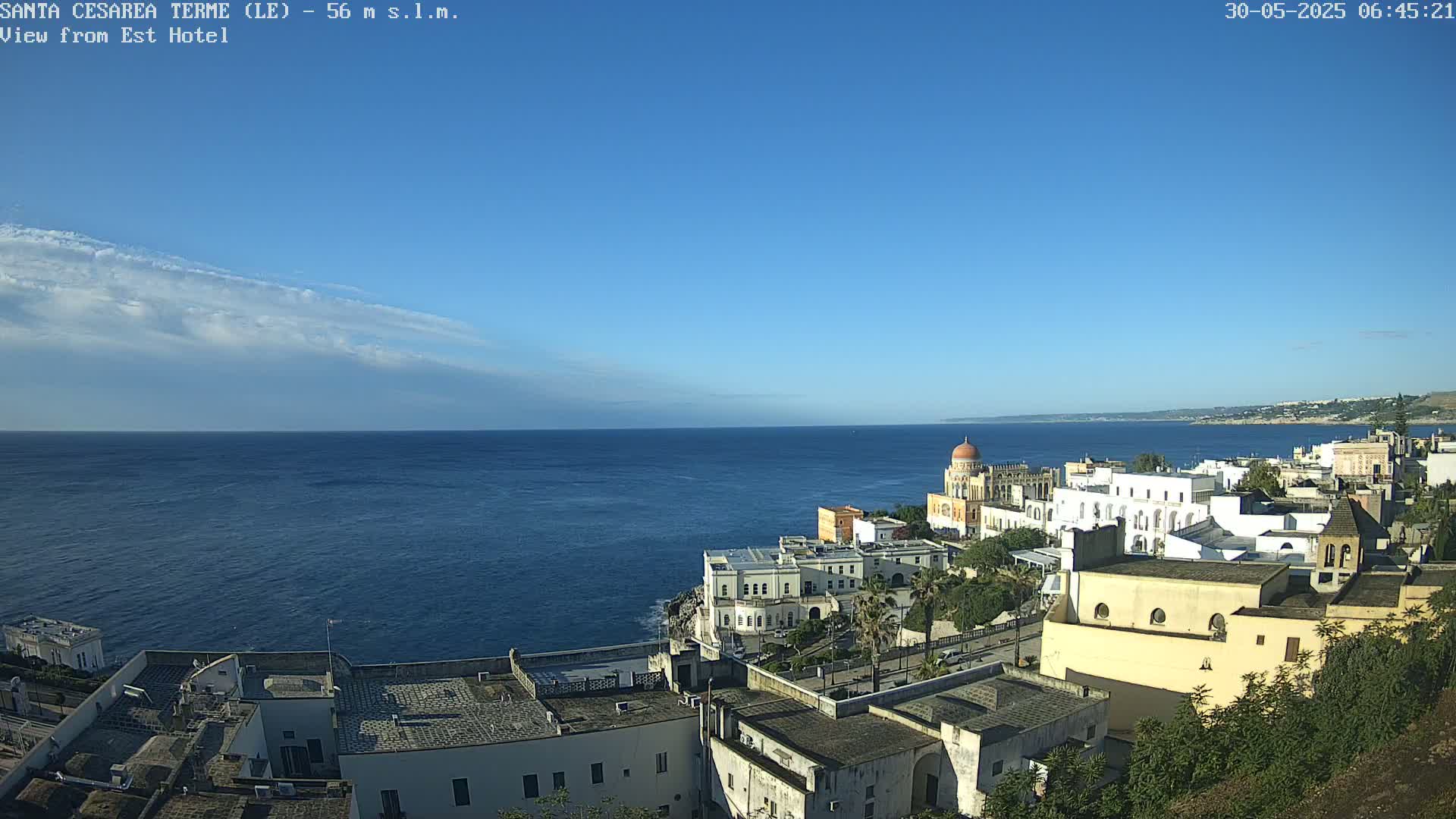 A coastal town with white buildings and a domed structure overlooks a calm, blue sea under a mostly clear, bright sky with a thin layer of clouds.