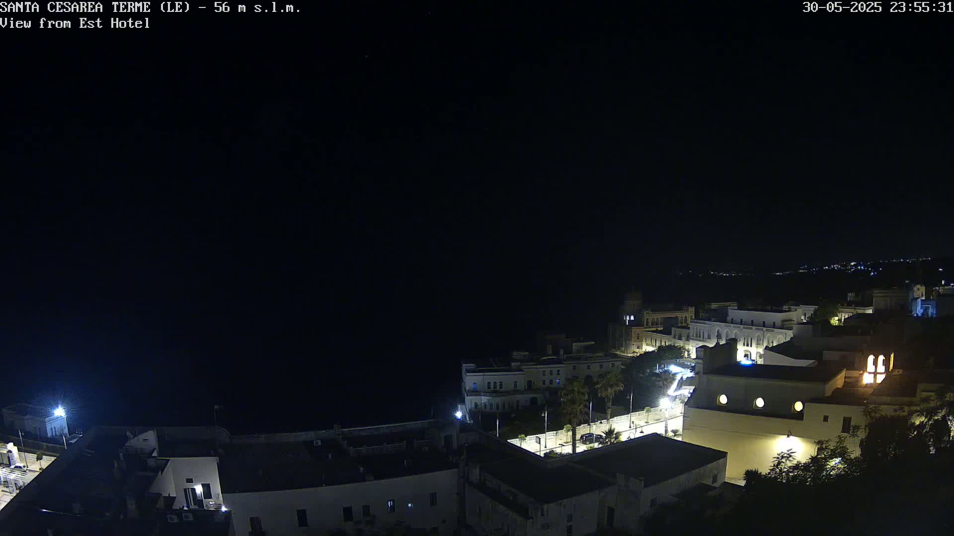 A nighttime, overhead view of a coastal town with buildings lit up, overlooking a dark sea under a clear sky.