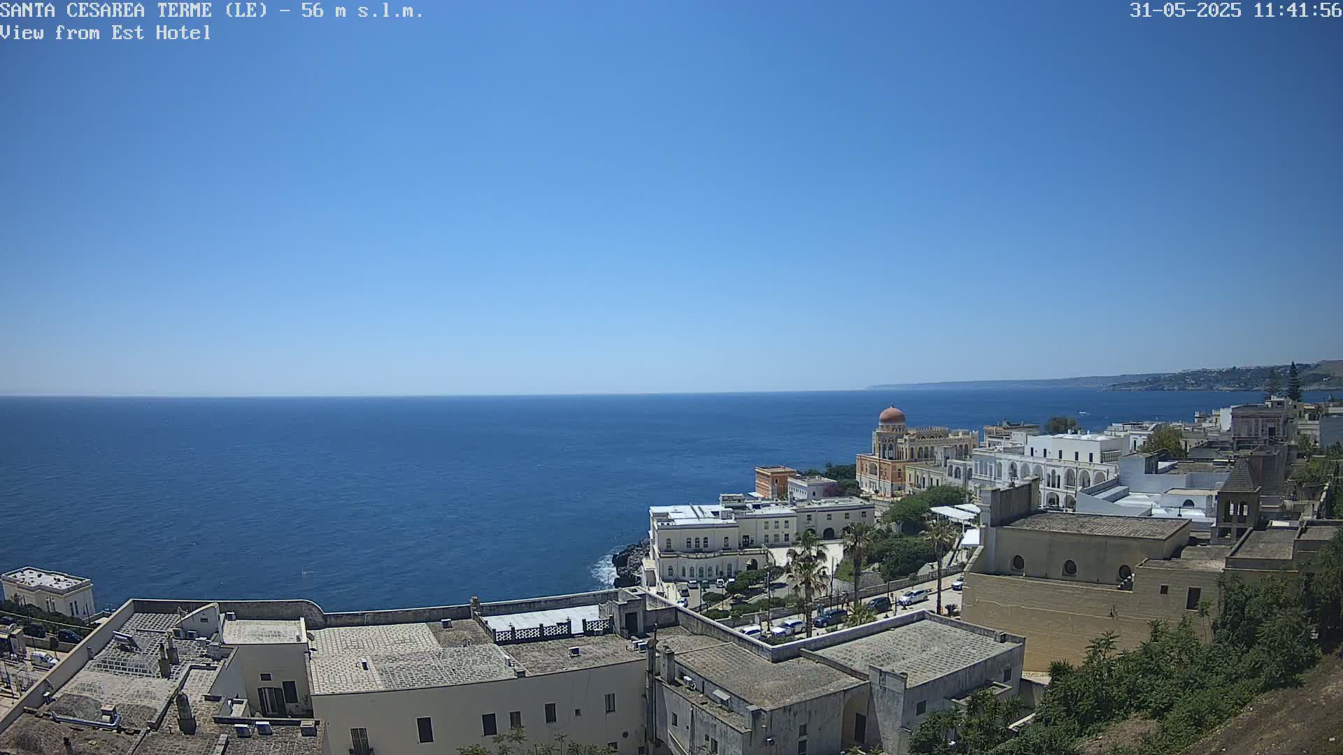 A coastal town with white buildings and a domed structure overlooks a calm, blue sea under a clear, sunny sky.