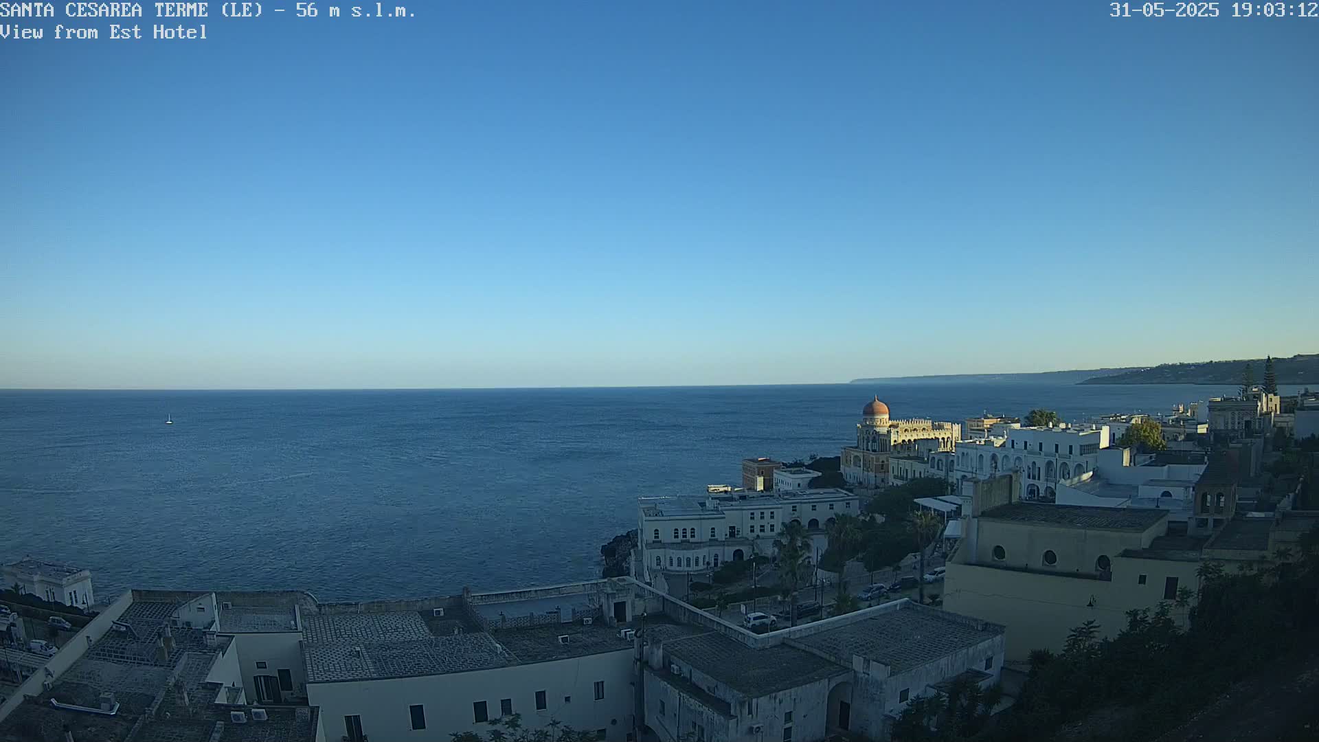 A coastal town with white buildings and a domed structure overlooks a calm, blue sea under a clear blue sky.