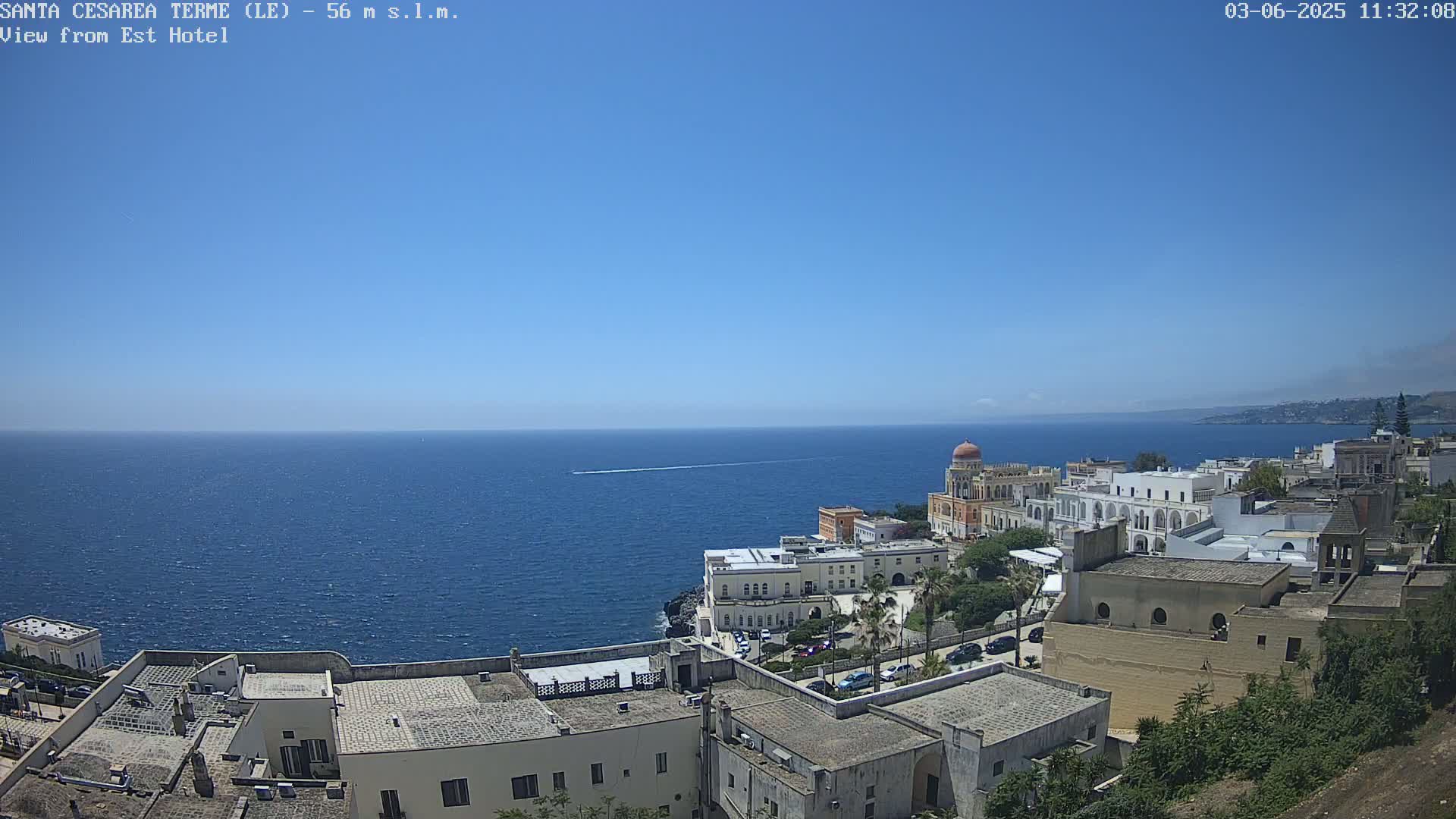 A coastal town with white buildings and a domed structure overlooks a calm, blue sea under a clear blue sky.