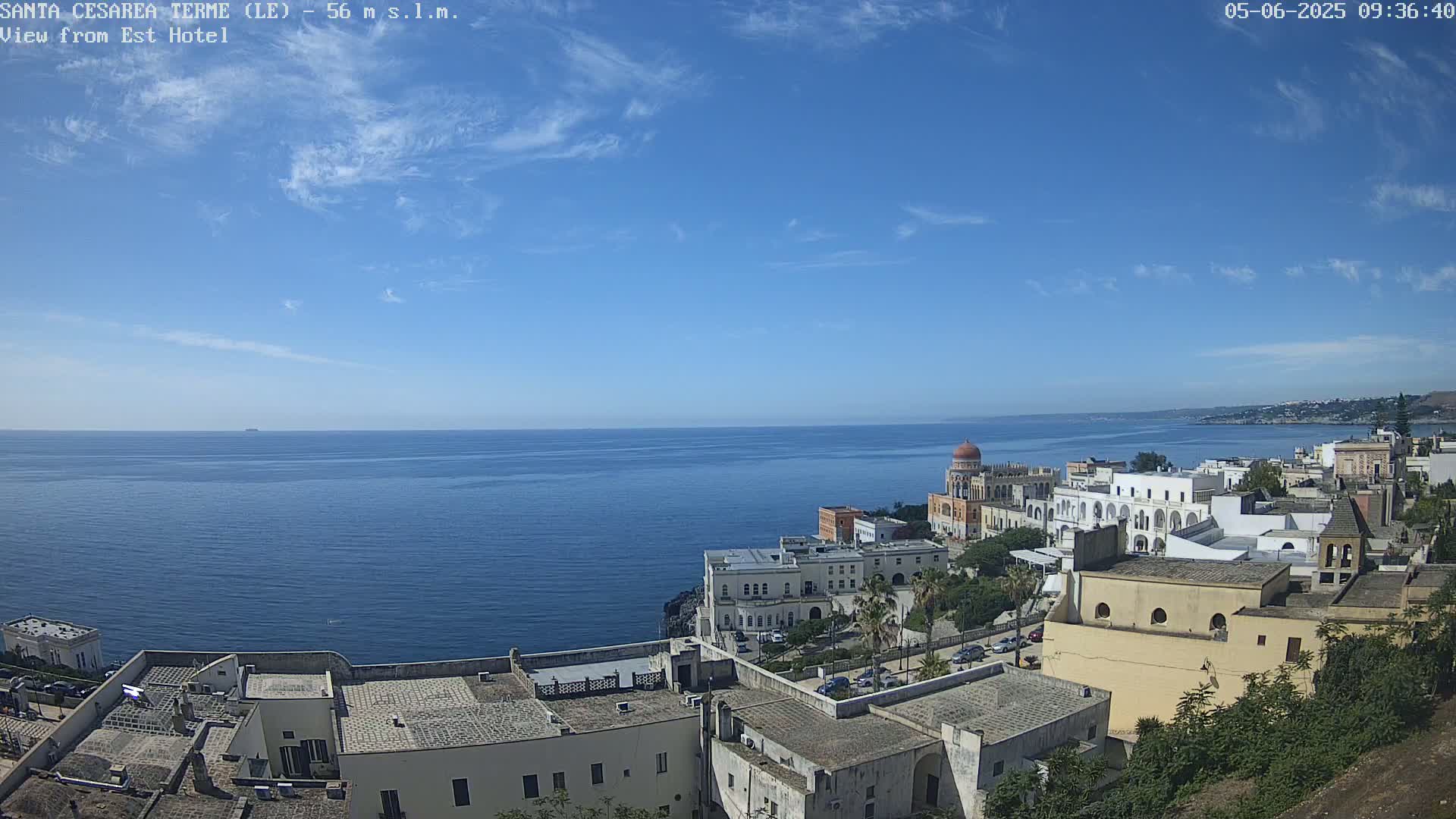 A coastal town with white buildings and a domed structure overlooks a calm blue sea under a mostly clear blue sky with a few scattered clouds.