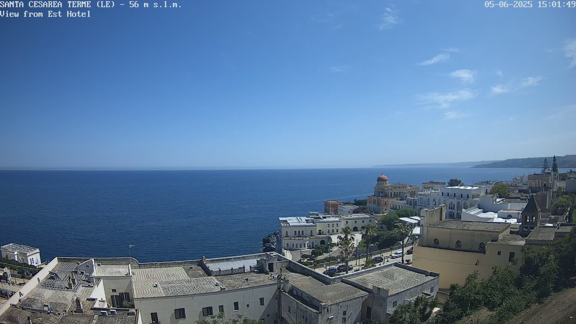 A coastal town with white buildings and a domed structure overlooks a calm, blue sea under a clear, sunny sky.