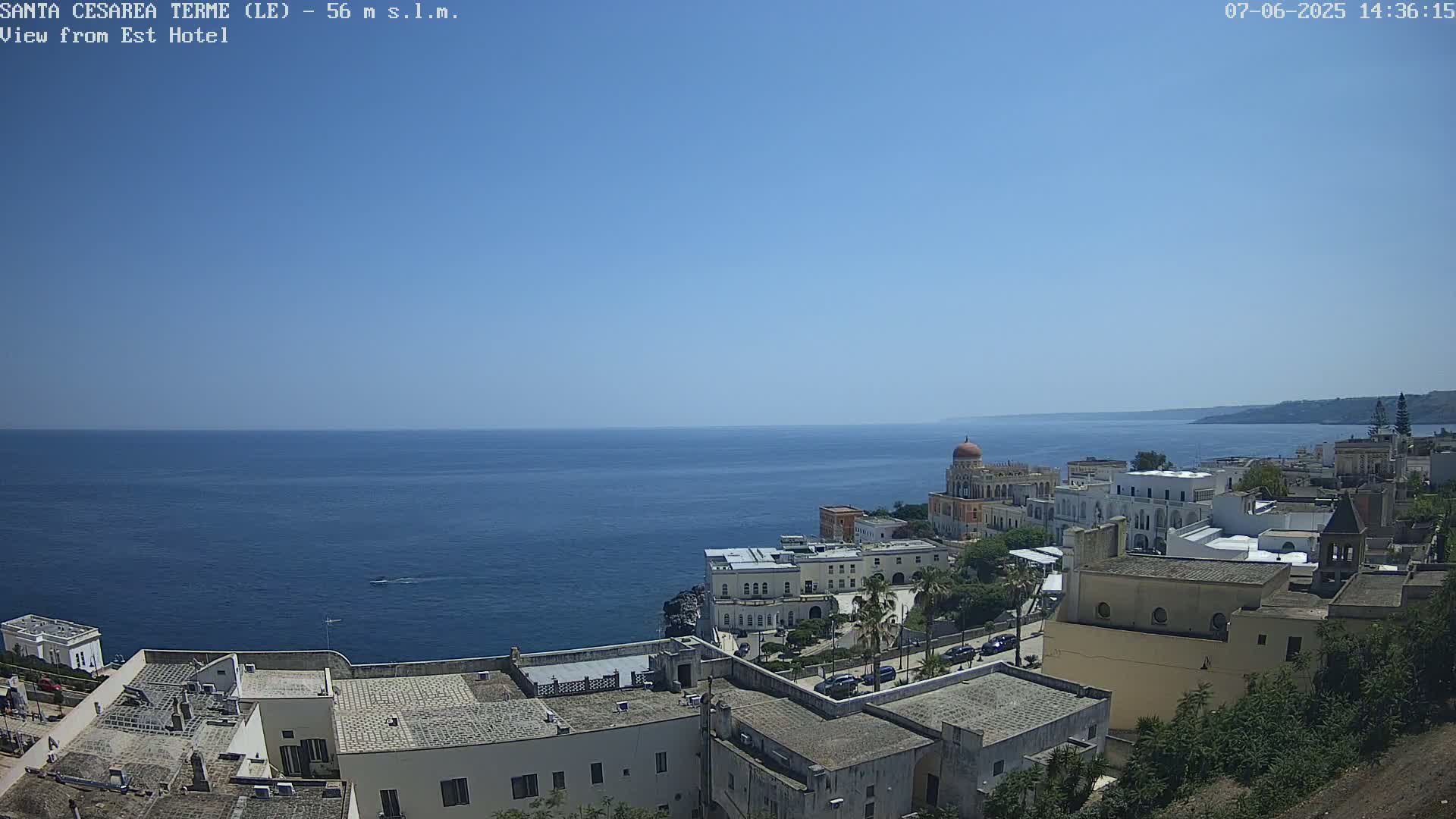 A coastal town with white buildings and a domed structure overlooks a calm, blue sea under a clear, sunny sky.