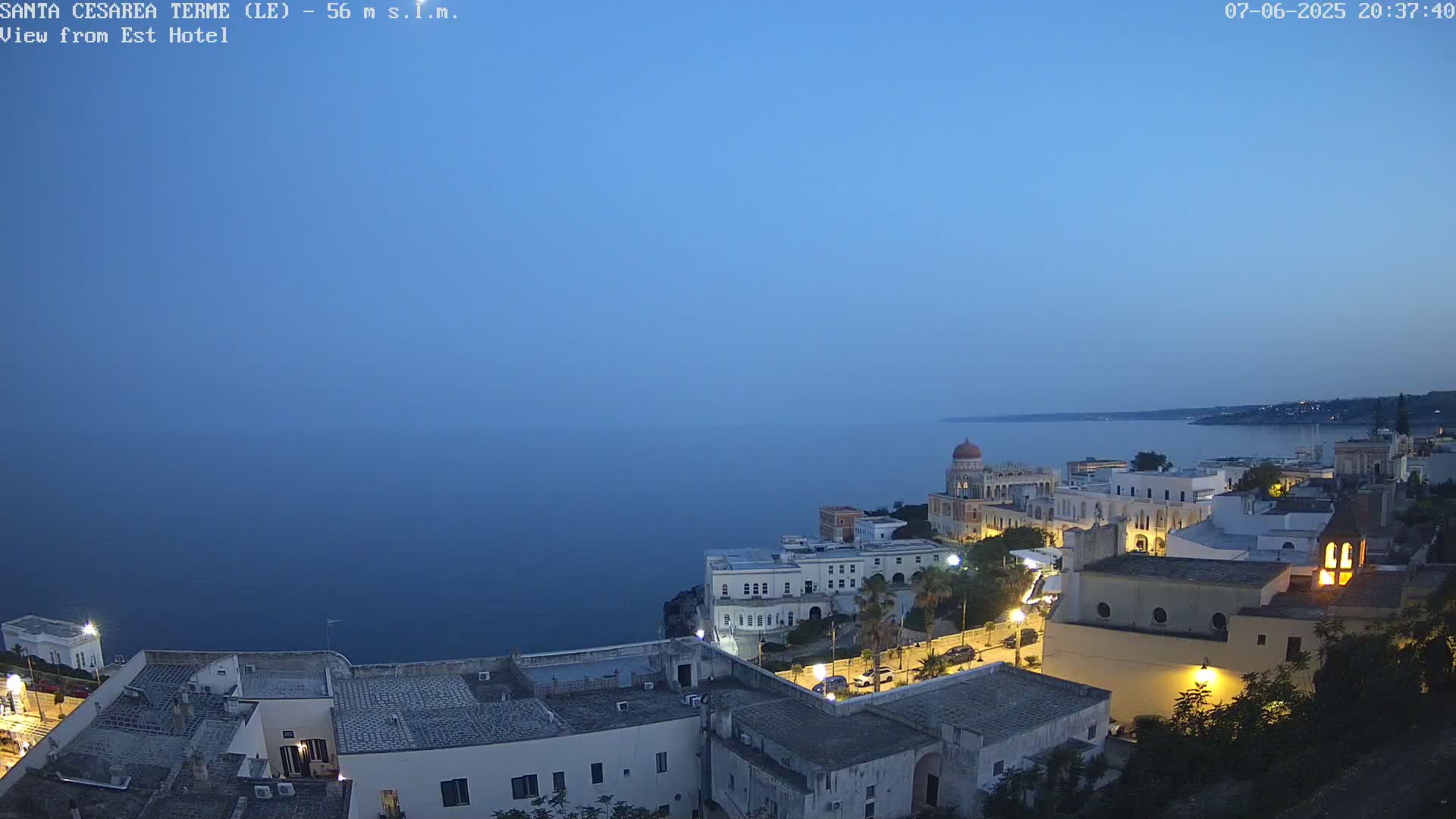 A coastal town at twilight, with calm, dark-blue water extending to the horizon under a clear, dusky sky.