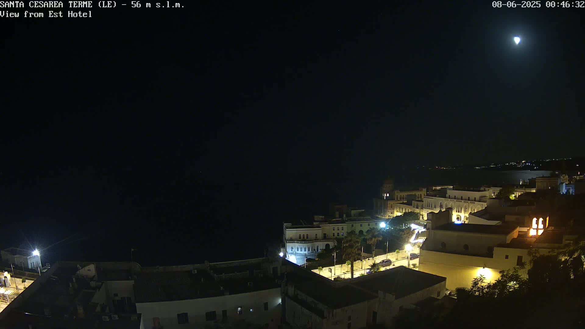 A nighttime, high-angle view of a coastal town with buildings lit up under a clear sky and a bright moon.