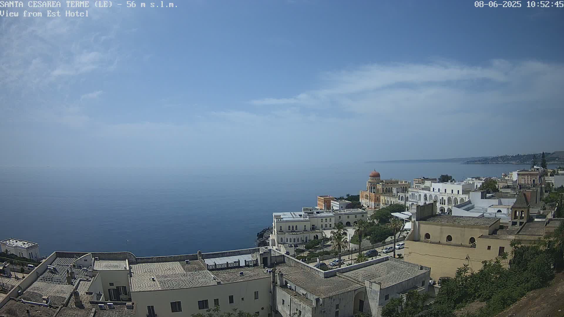 A coastal town with white buildings and a calm blue sea under a mostly clear sky with a few clouds.