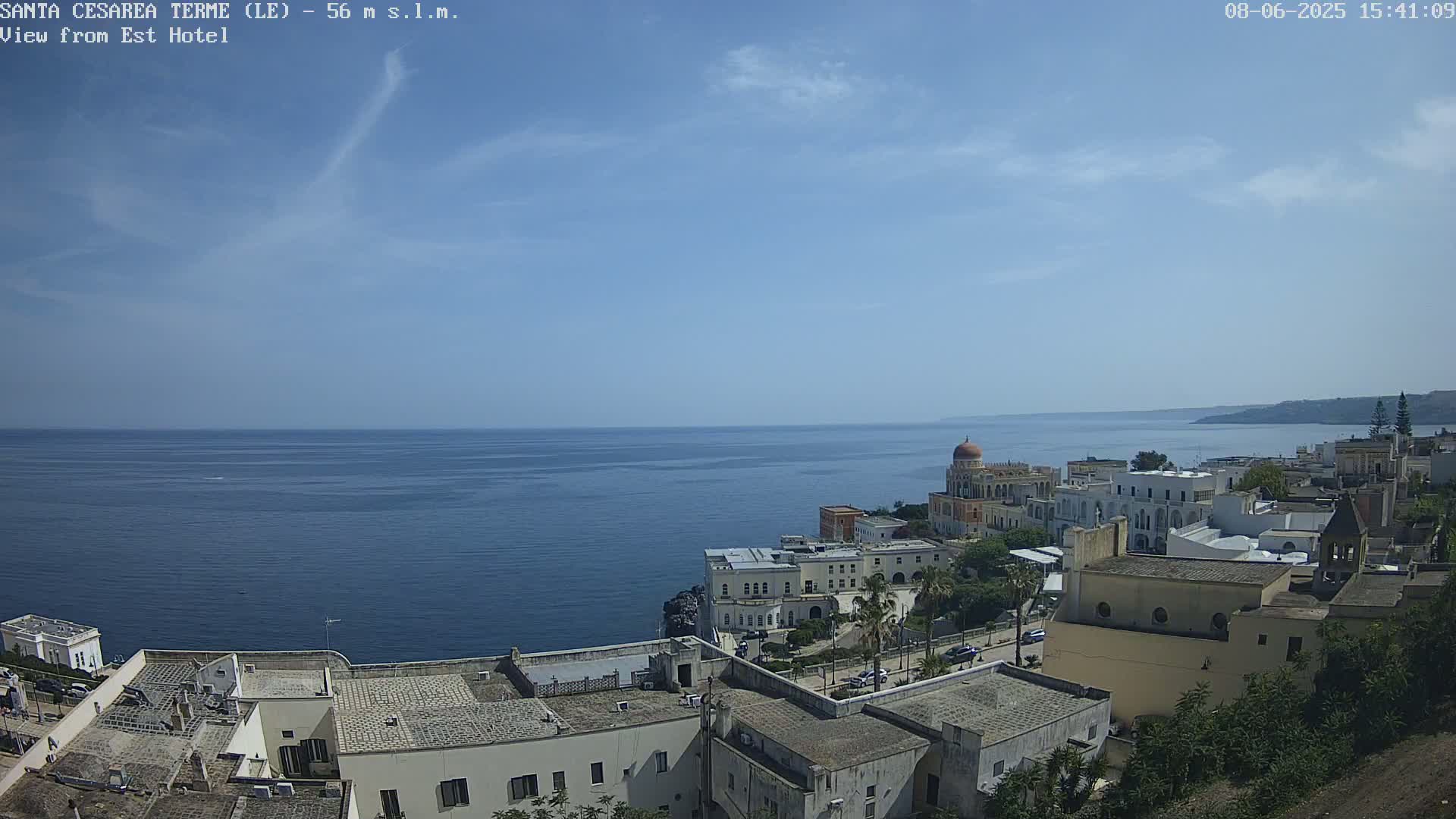 A coastal town with white buildings and a domed structure overlooks a calm blue sea under a mostly clear sky.