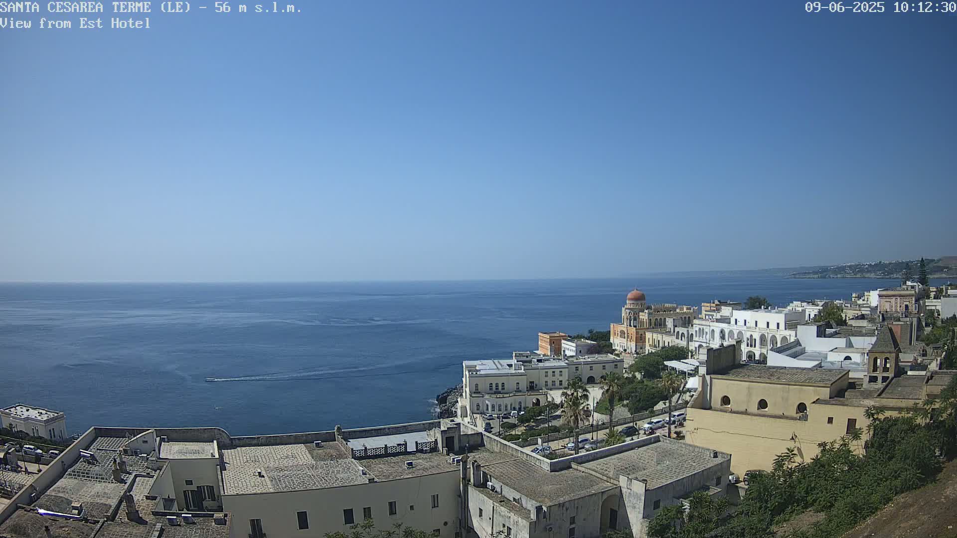 A coastal town with white buildings and a domed structure overlooks a calm, blue sea under a clear, sunny sky.