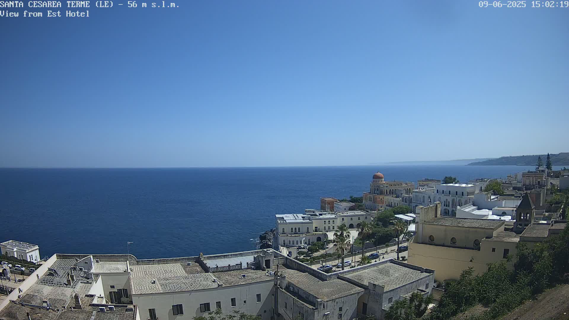 A coastal town with white buildings and a domed structure overlooks a calm, blue sea under a clear blue sky.