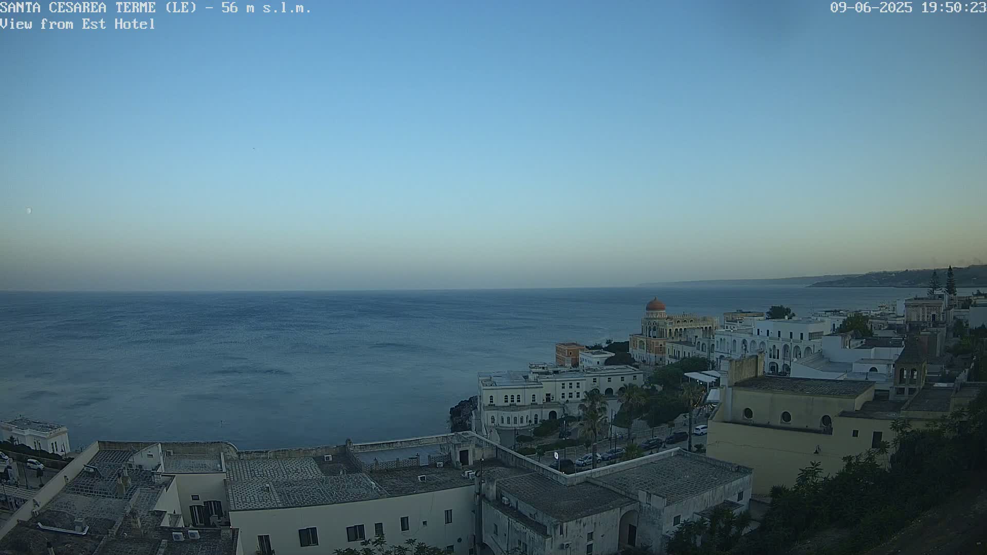 A coastal town with white buildings overlooks a calm, dark blue sea under a clear, twilight sky.