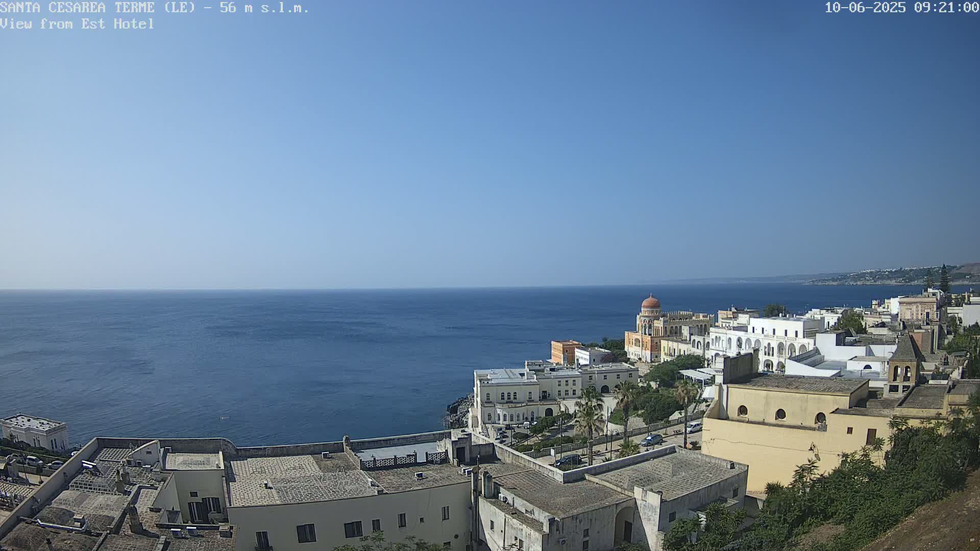 A coastal town with white buildings and a domed structure overlooks a calm, blue sea under a clear blue sky.