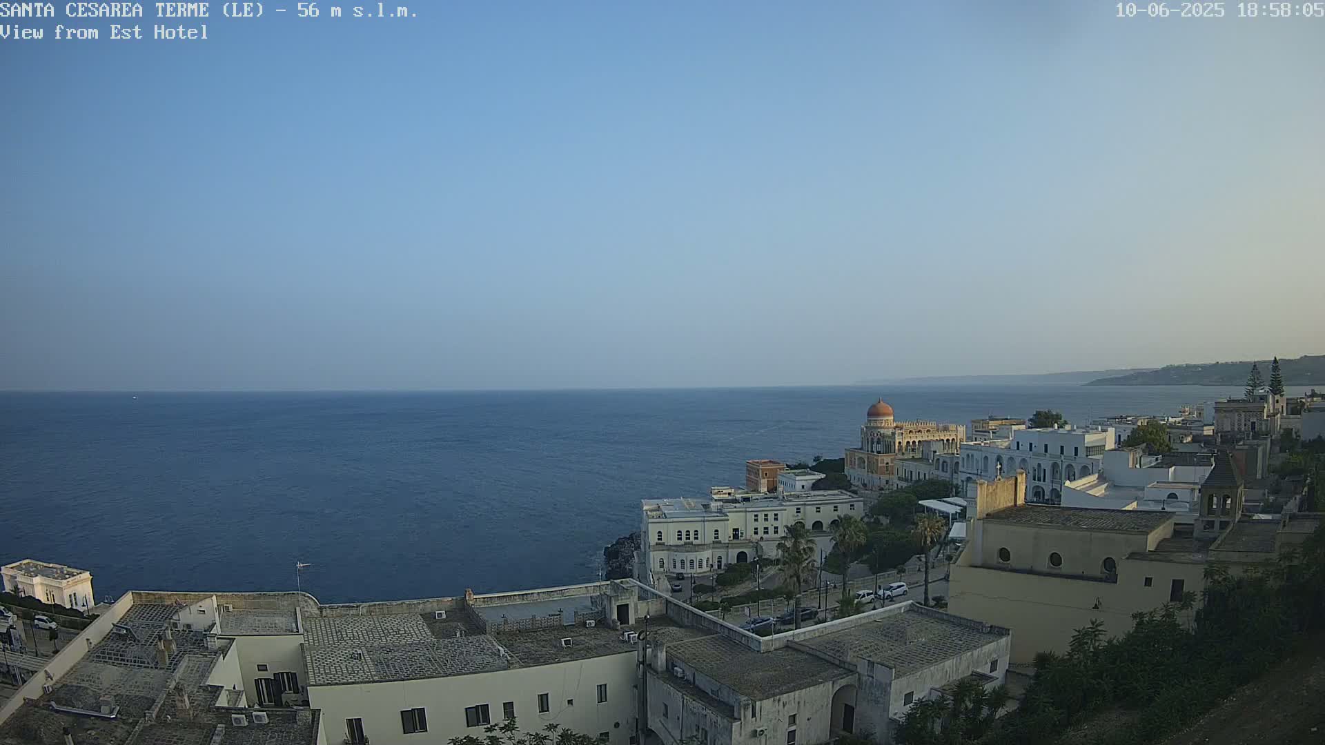 A coastal town with white buildings and a domed structure overlooks a calm, blue sea under a clear sky.