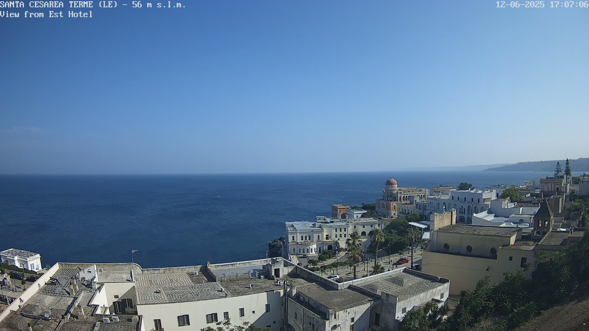 A coastal town with white buildings and a domed structure overlooks a calm, blue sea under a clear, sunny sky.