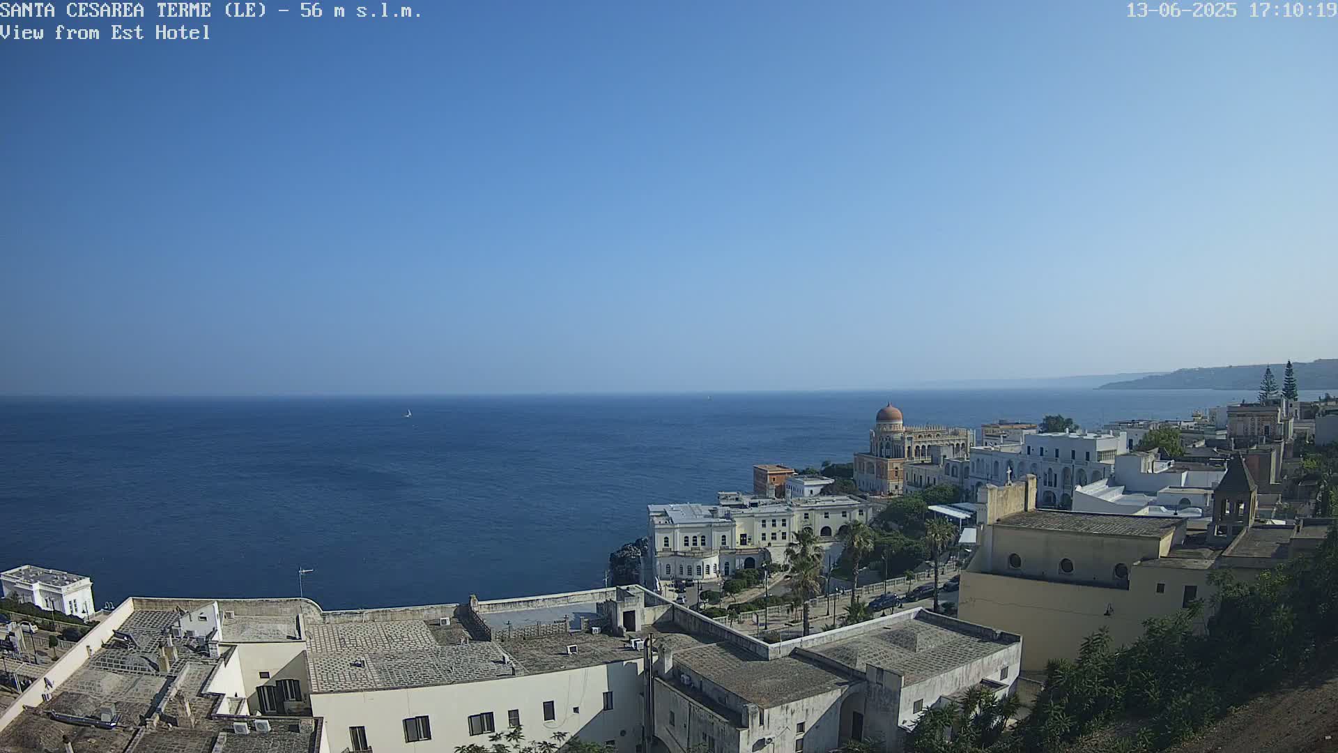 A coastal town with white buildings and a domed structure overlooks a calm, blue sea under a clear sky.
