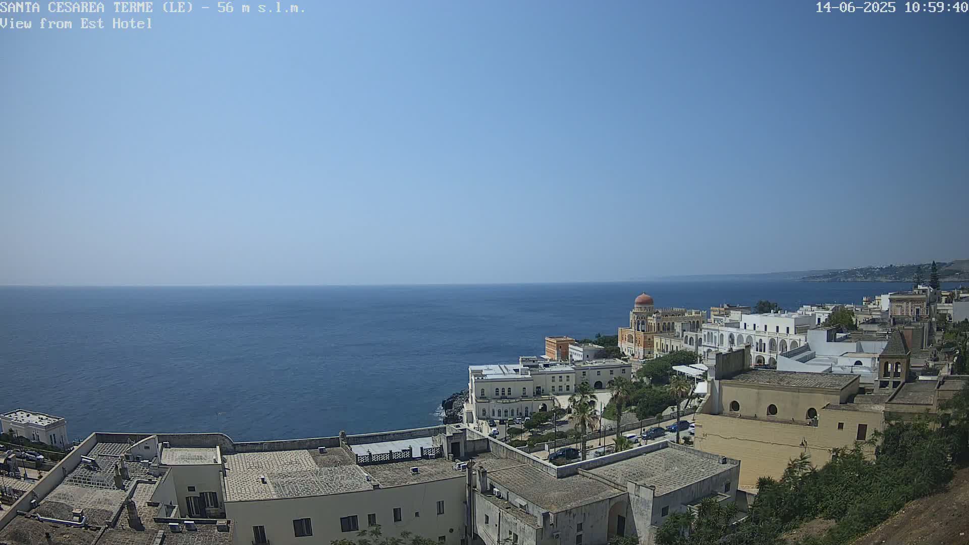 A coastal town with white buildings and a domed structure overlooks a calm blue sea under a clear sky.