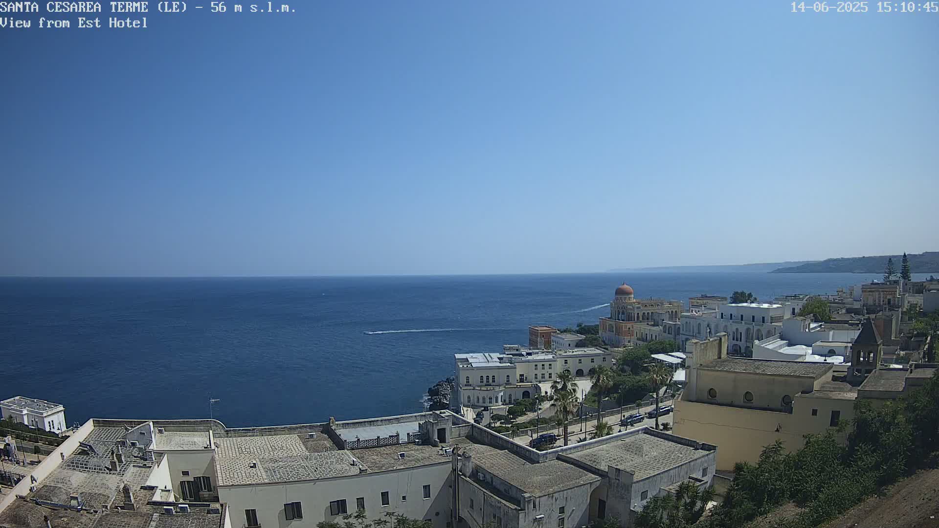 A coastal town with white buildings and a calm blue sea under a clear blue sky is seen from an elevated vantage point.