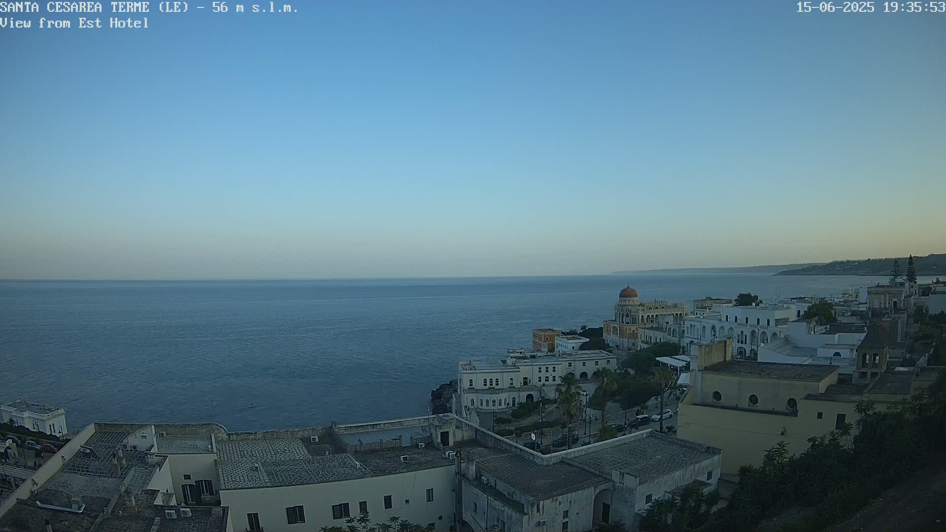 A coastal town with white buildings and a domed structure sits on a cliff overlooking a calm, dark-blue sea under a clear, light-blue sky.