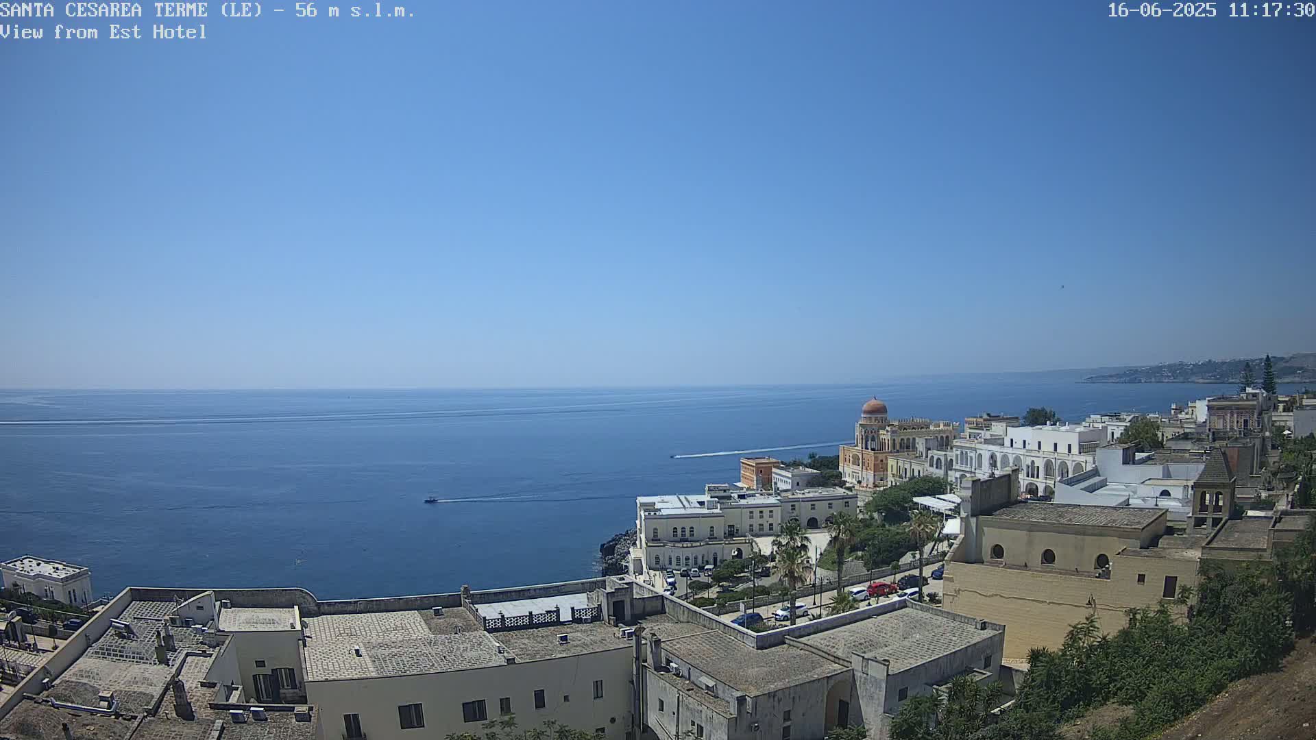 A coastal town with white buildings and a domed structure overlooks a calm blue sea under a clear blue sky.