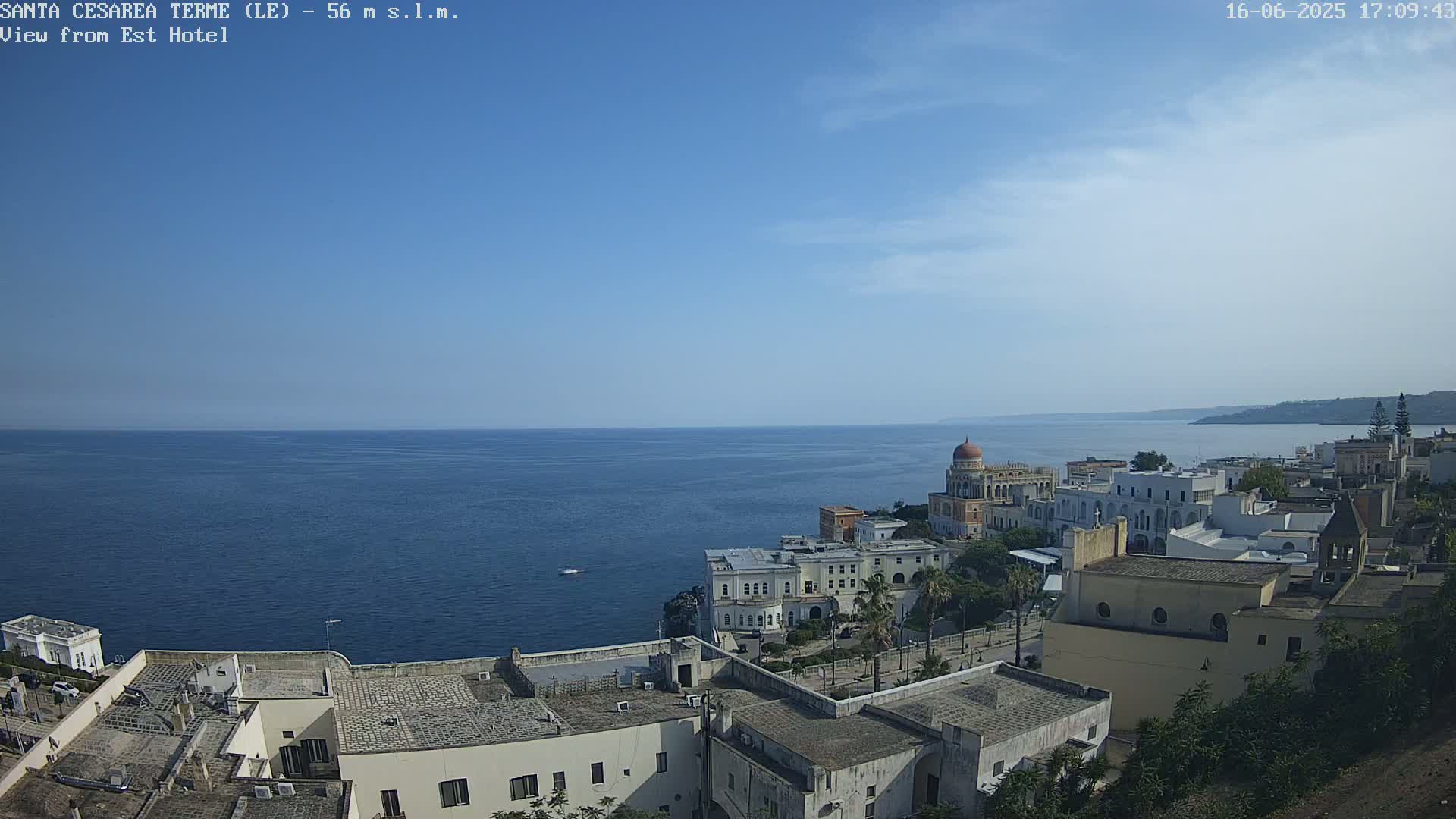 A coastal town with white buildings and a domed structure overlooks a calm blue sea under a clear sky.