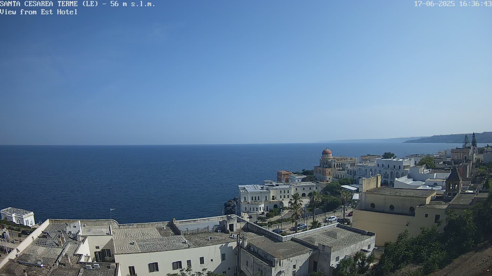 A coastal town with white buildings overlooks a calm, blue sea under a clear, sunny sky.