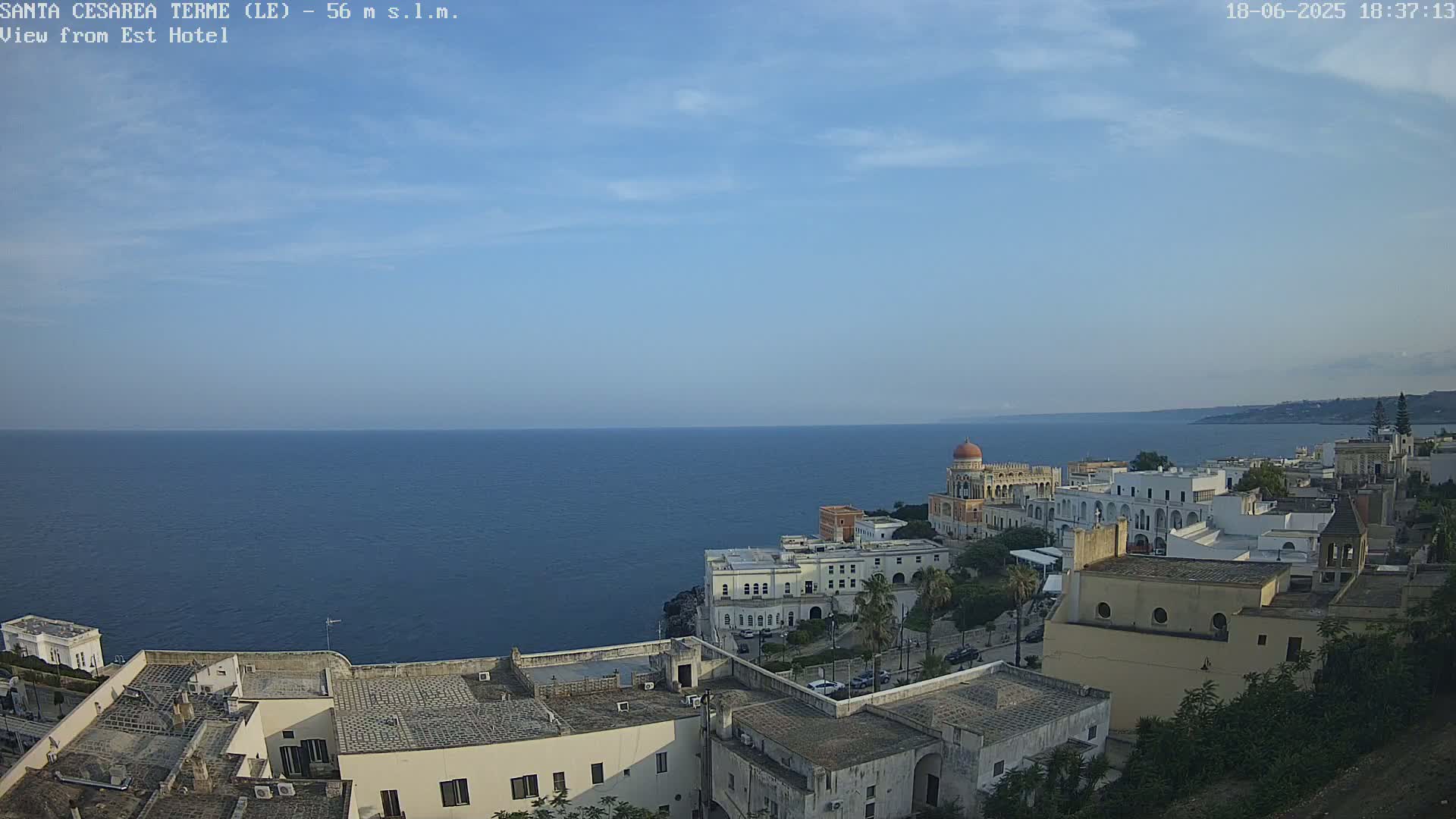 A coastal town with white buildings, a prominent domed structure, and a calm blue sea under a mostly clear sky.
