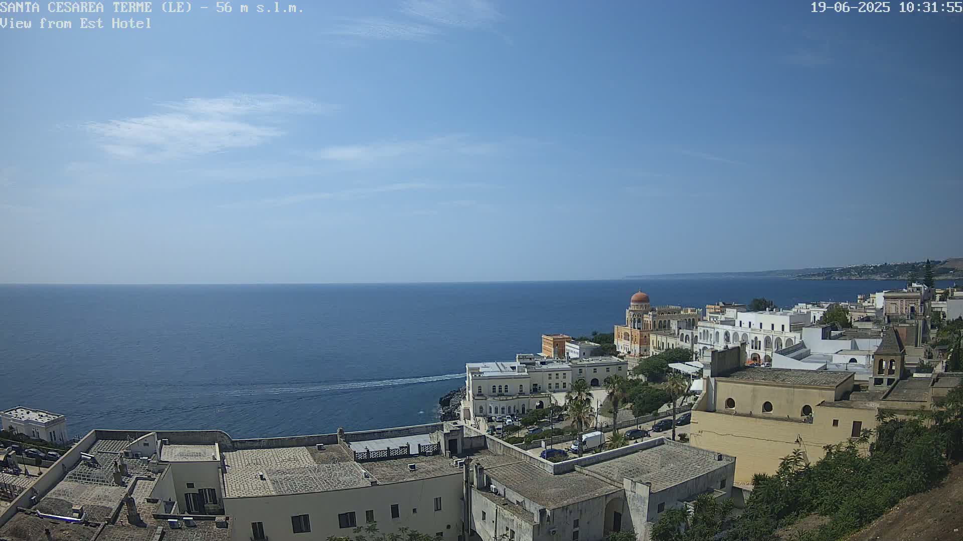 A coastal town with white buildings and a domed structure overlooks a calm blue sea under a clear sky.