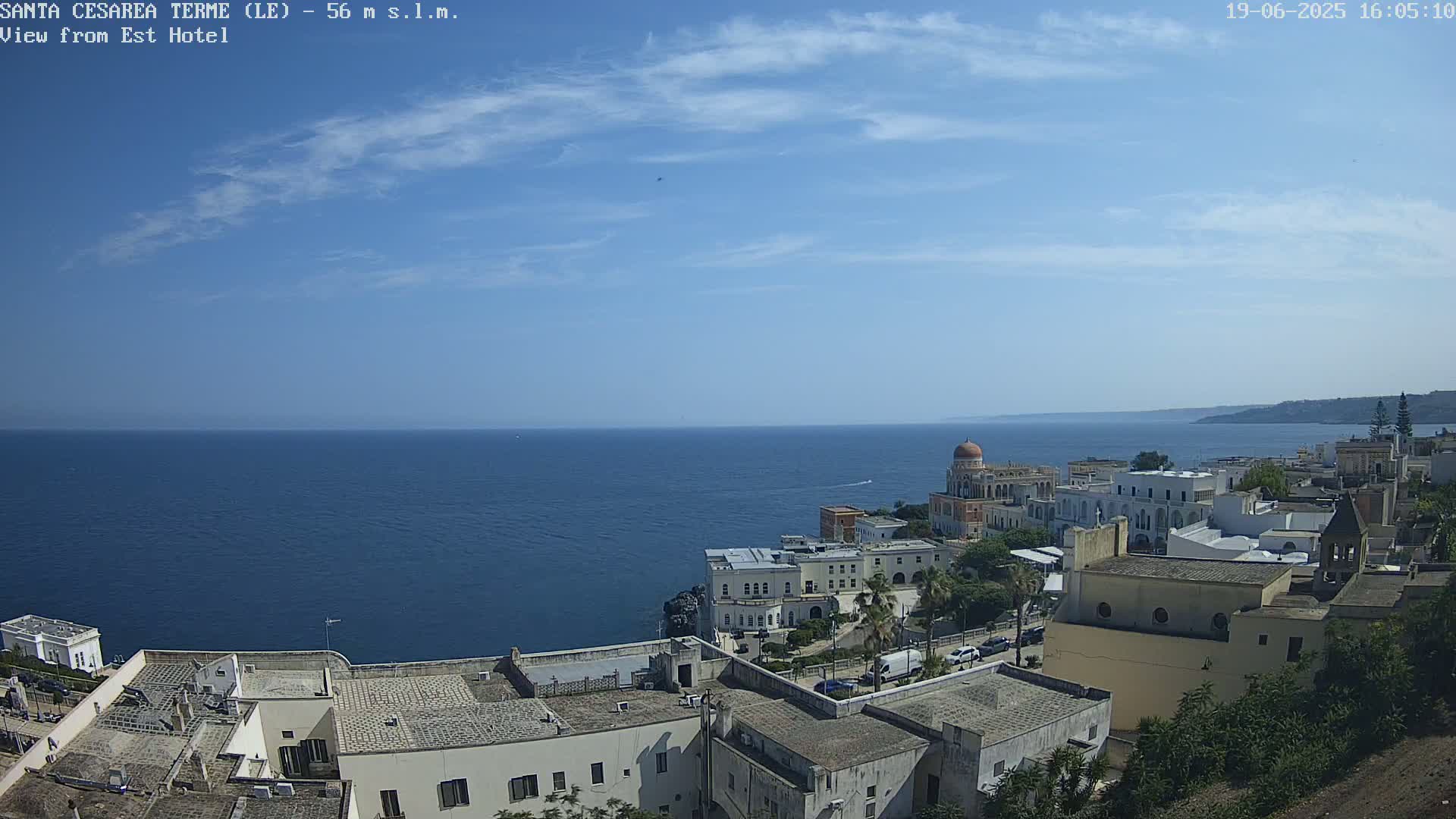A coastal town with white buildings, a domed structure, and lush greenery overlooks a calm, blue sea under a mostly sunny sky.