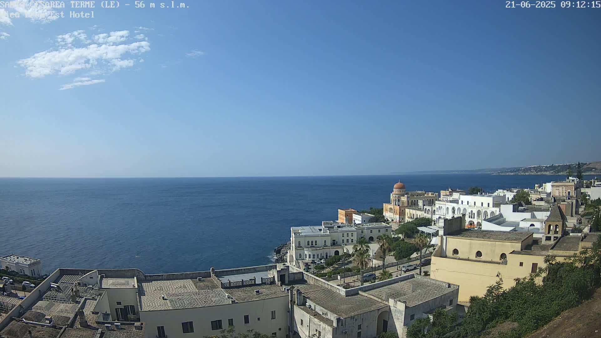 A coastal town with white buildings overlooks a calm, blue sea under a clear, sunny sky.