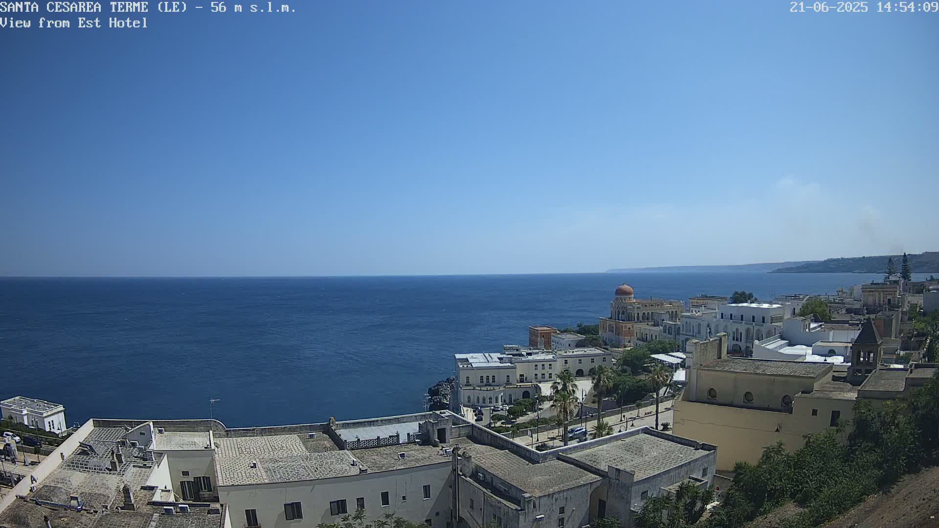 A coastal town with white buildings sits on a cliff overlooking a calm, deep blue sea under a clear, bright blue sky.