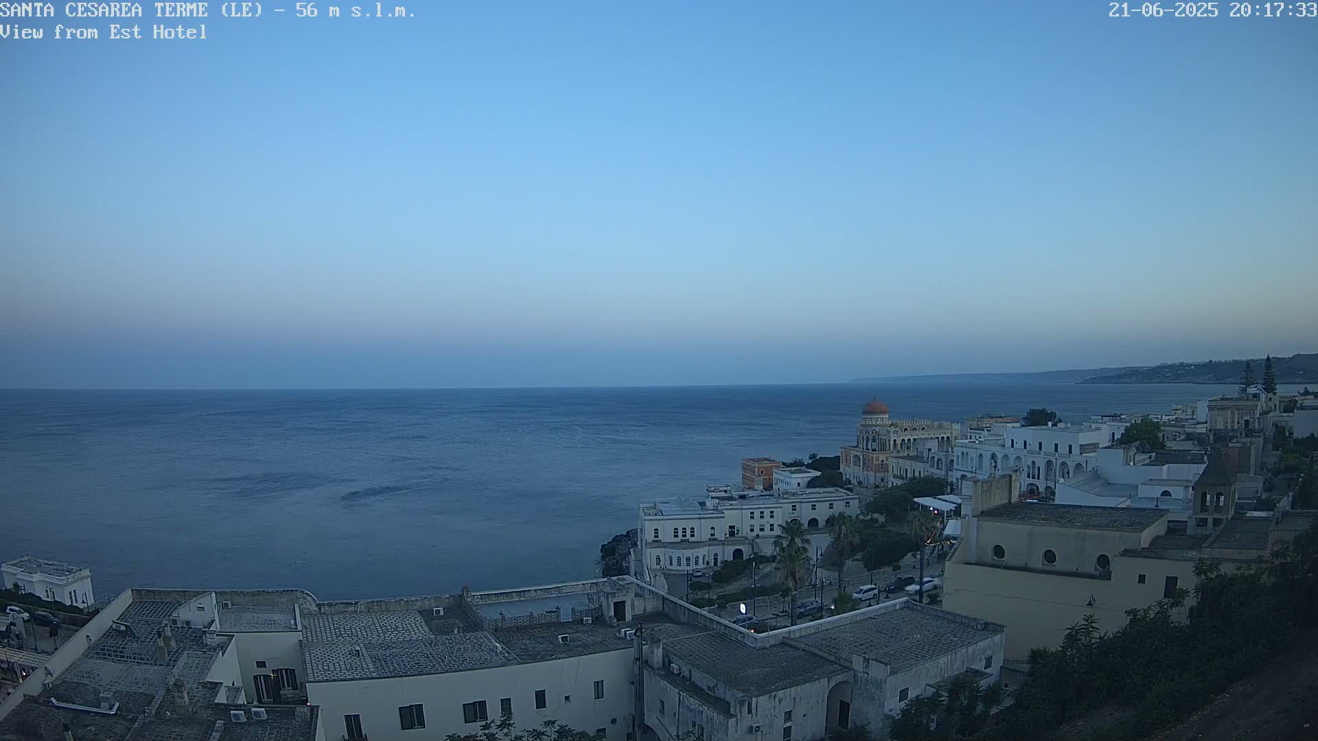 A coastal town with white buildings overlooks a calm, dark-blue sea under a twilight sky.