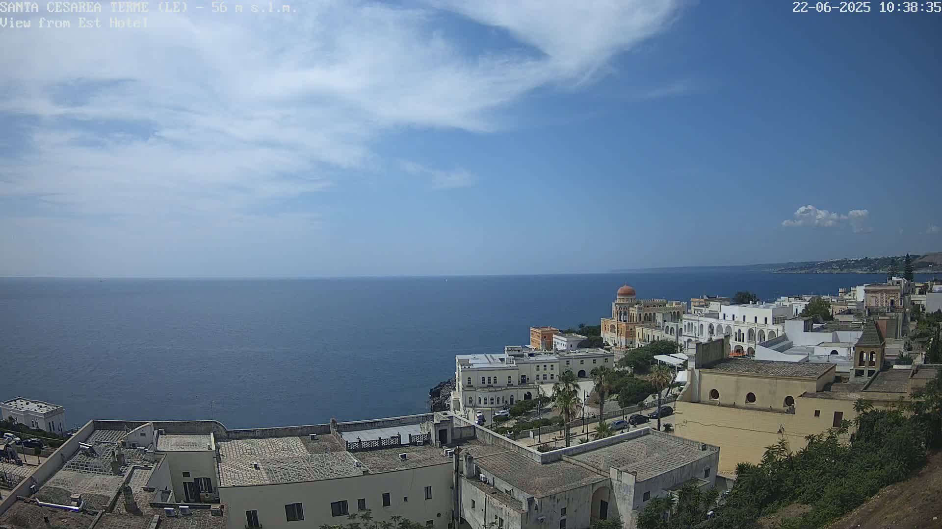 A coastal town with white buildings and a domed structure overlooks a calm blue sea under a mostly clear, sunny sky.