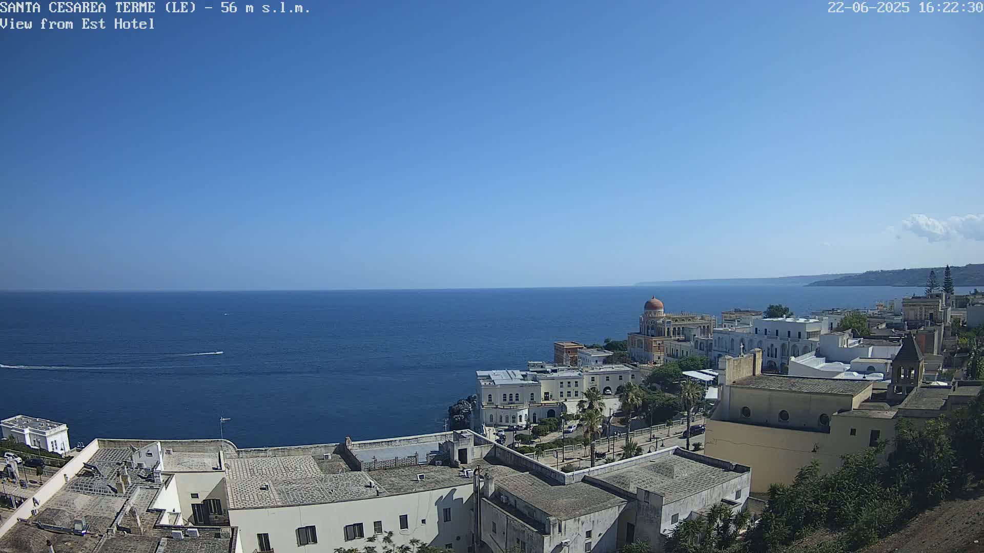 A coastal town with white buildings and a domed structure overlooks a calm, blue sea under a clear, sunny sky.
