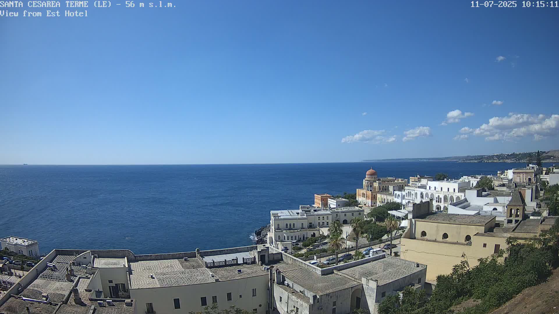 A coastal town with white buildings and a dome-topped structure overlooks a calm blue sea under a mostly clear, sunny sky with a few scattered clouds.