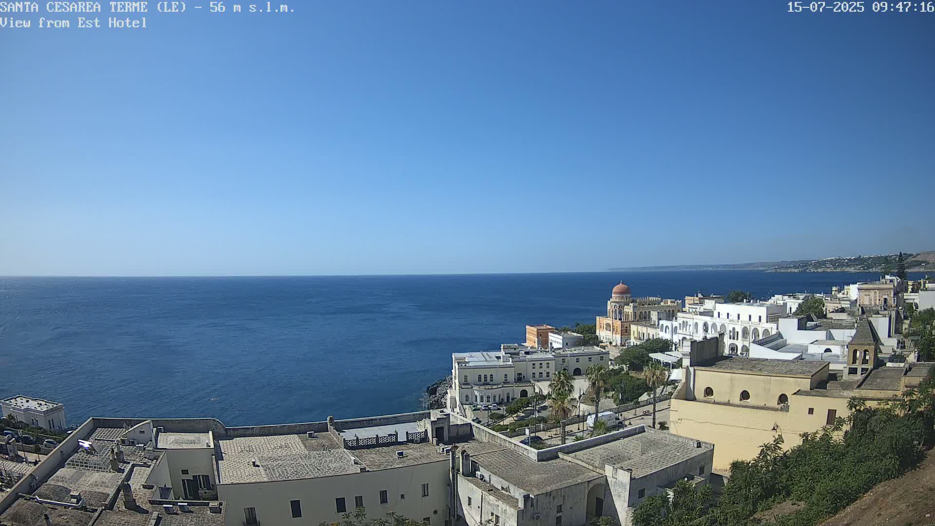 A coastal town with white buildings, a prominent domed structure, and green vegetation overlooks a calm blue sea under a clear blue sky.